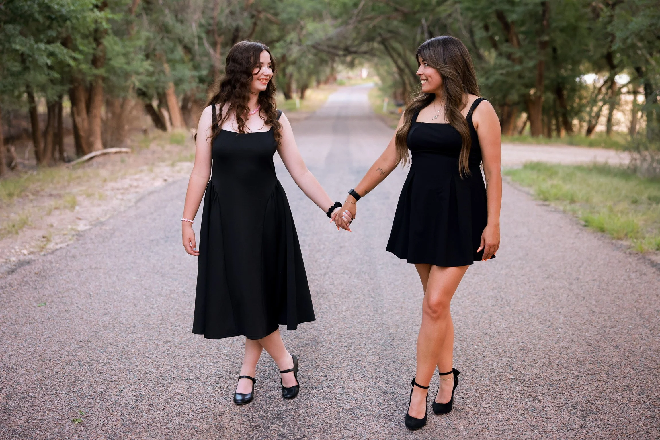 Two women in black dresses holding hands and smiling at each other on a gravel path surrounded by green trees.