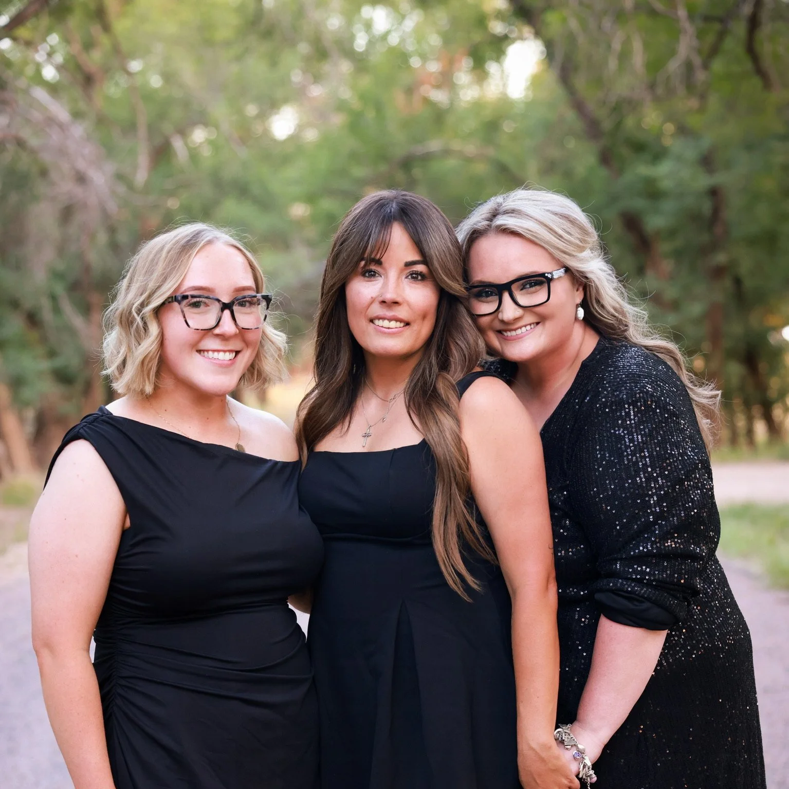 Three women standing outdoors in a wooded area, dressed in black dresses and wearing glasses, smiling at the camera.