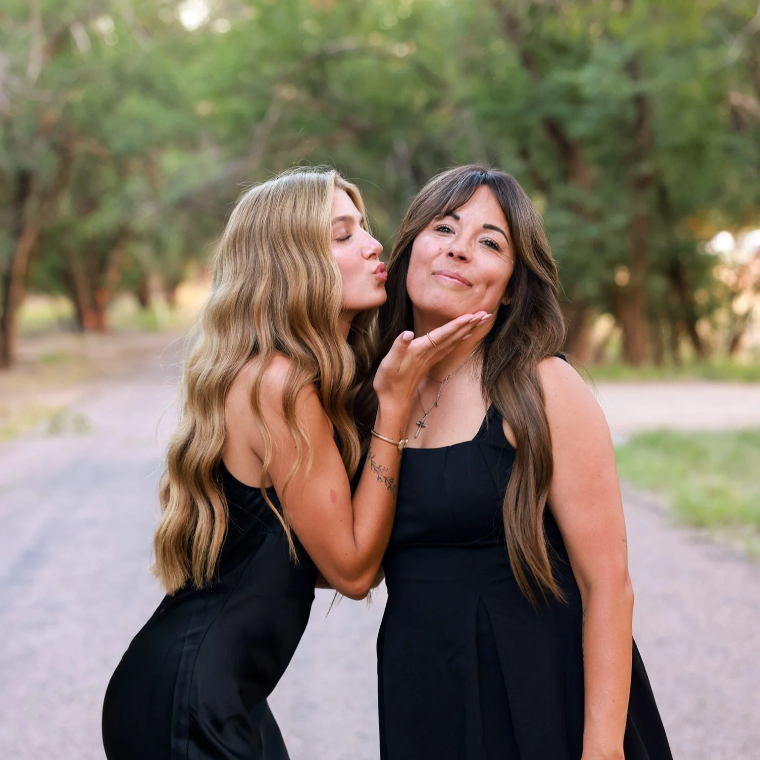 Two women in black dresses sharing a kiss in a park with green trees in the background.
