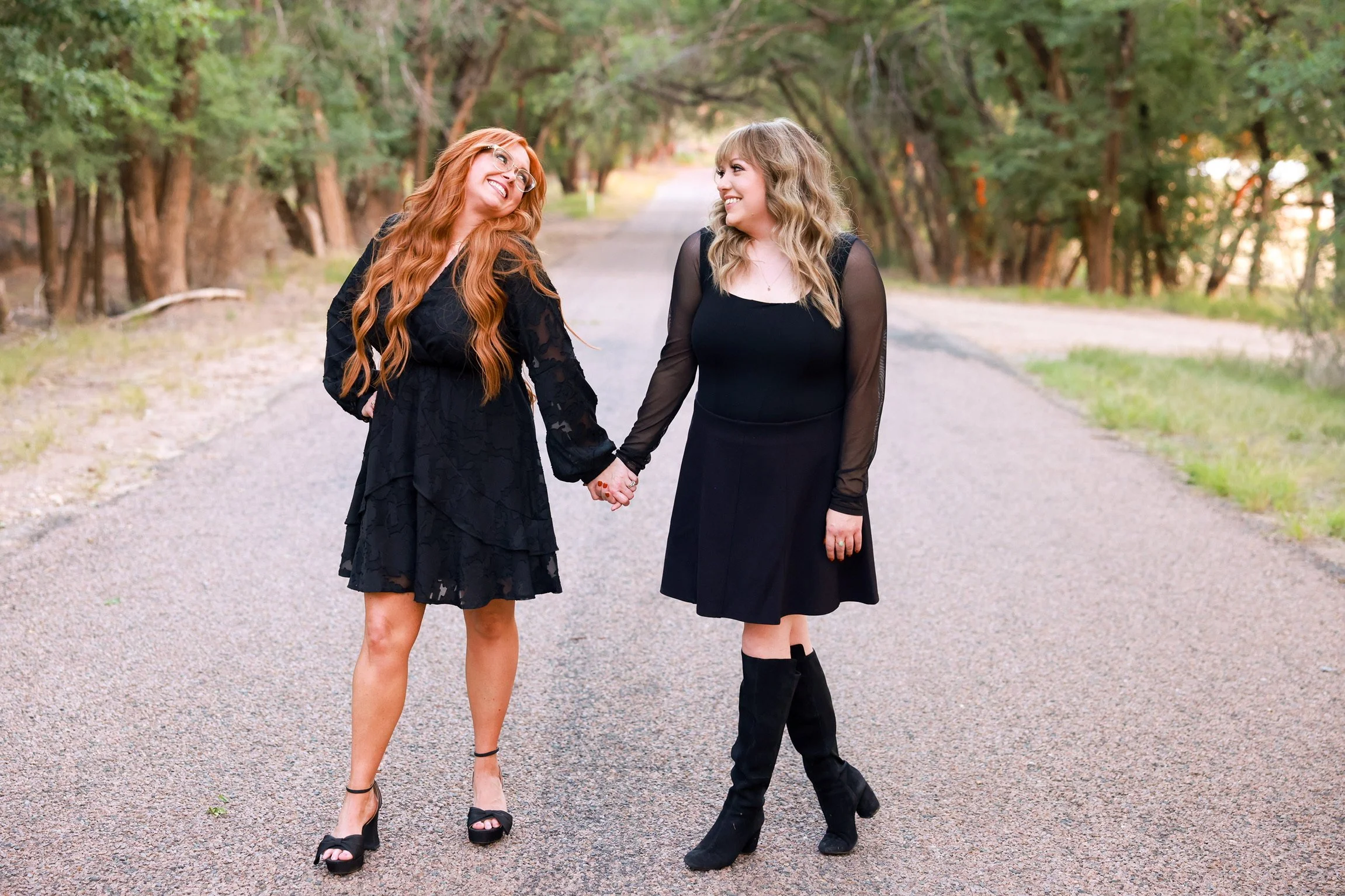 Two women holding hands and smiling at each other on a tree-lined gravel path, both dressed in black.