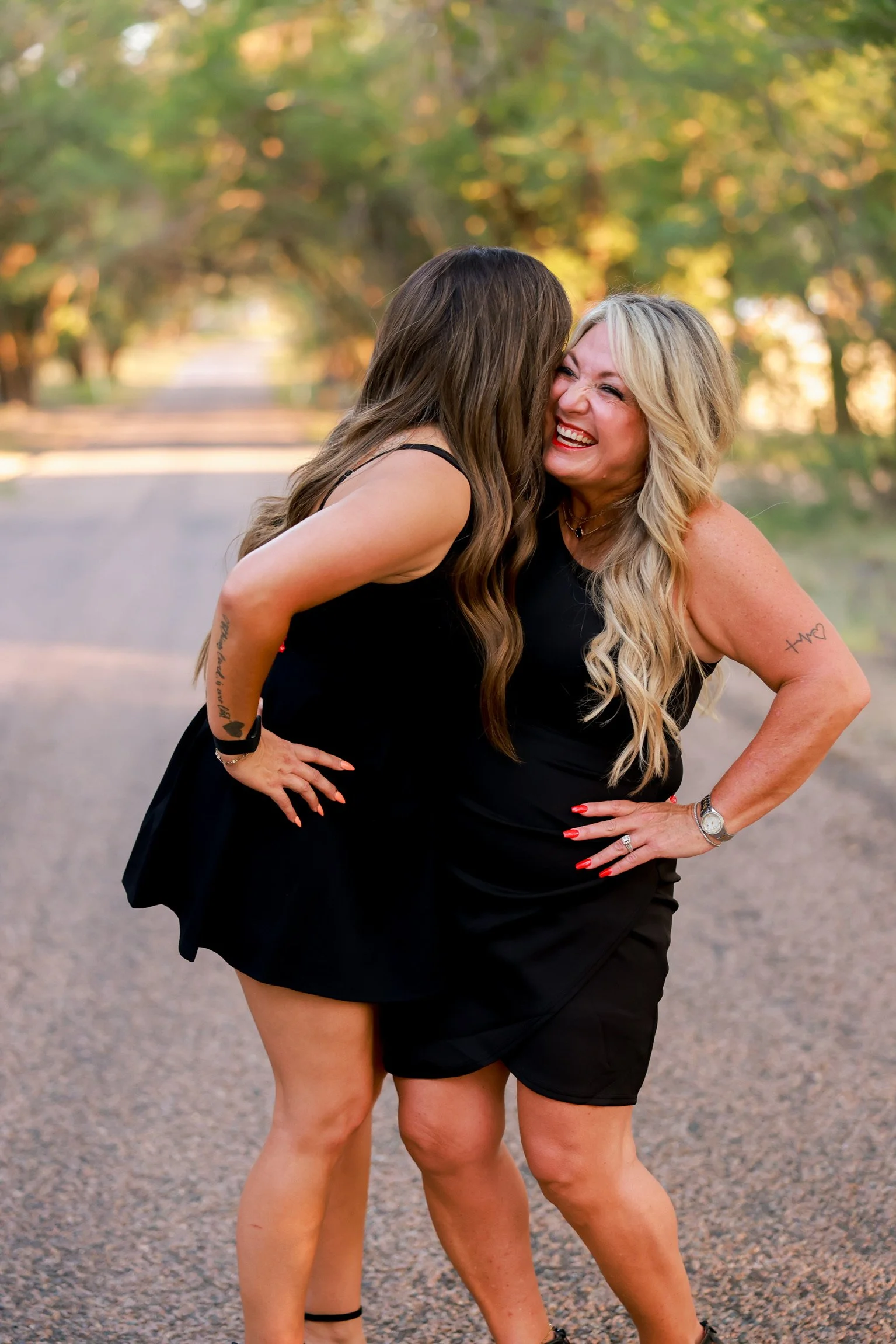 Two women wearing black dresses laughing and embracing each other outdoors on a sunny day, with trees and a gravel path in the background.