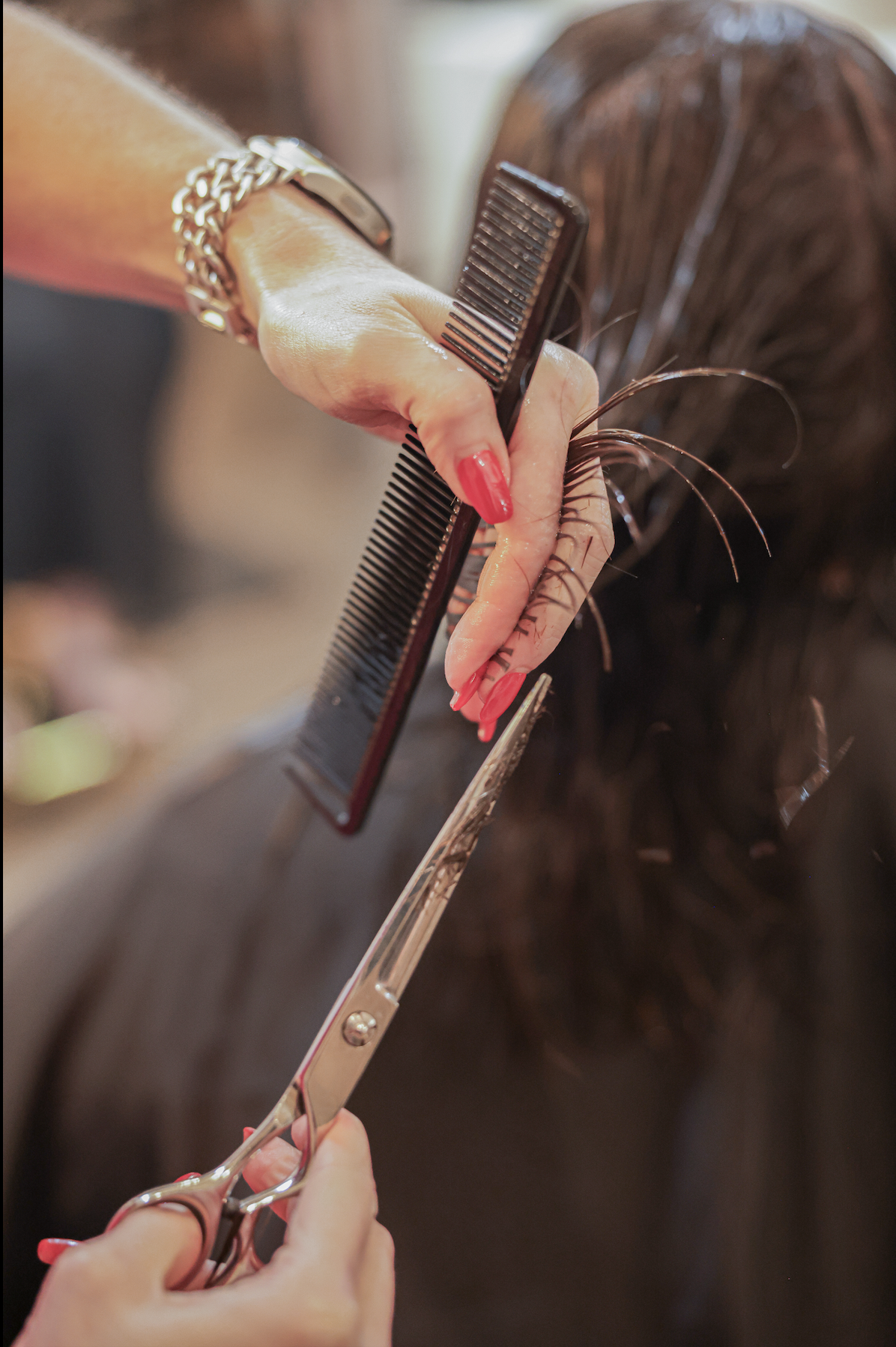 A person with painted red nails is cutting a person's brown hair with scissors and a comb.