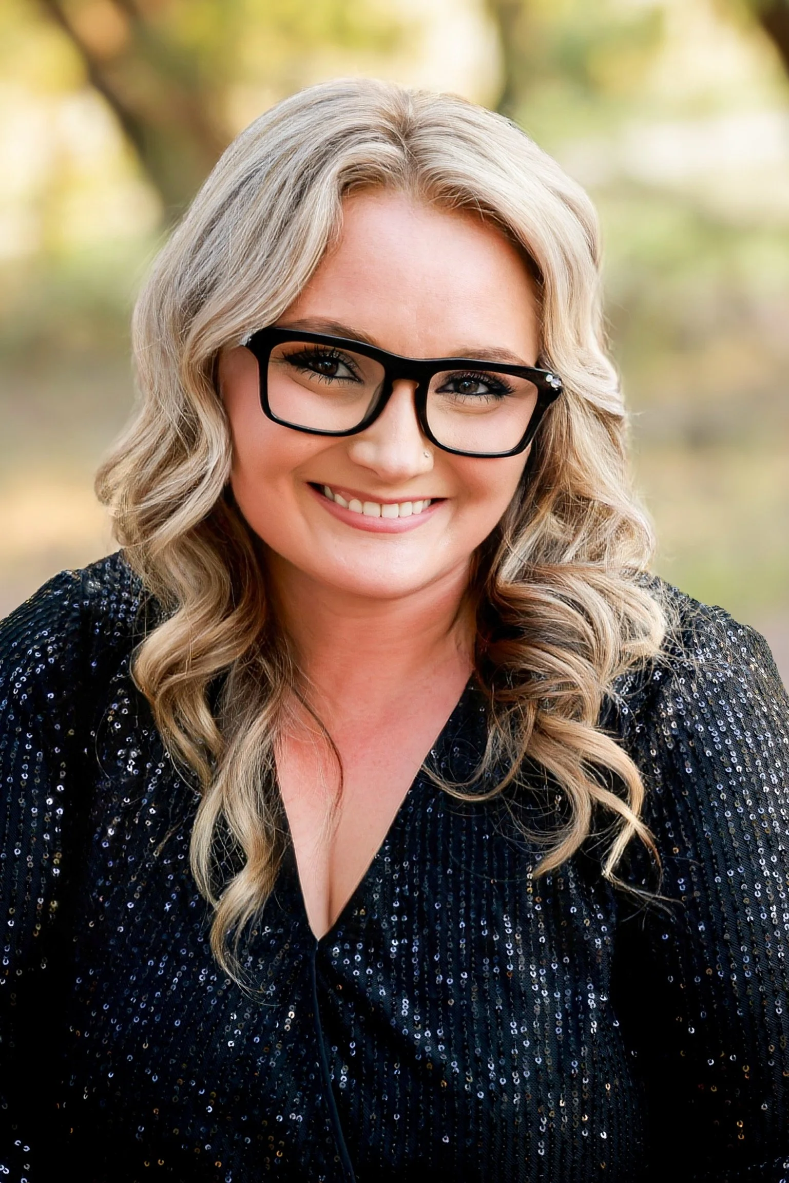 A smiling woman with blonde, curly hair wearing black-rimmed glasses and a black sequined top, outdoors with blurred greenery in the background.