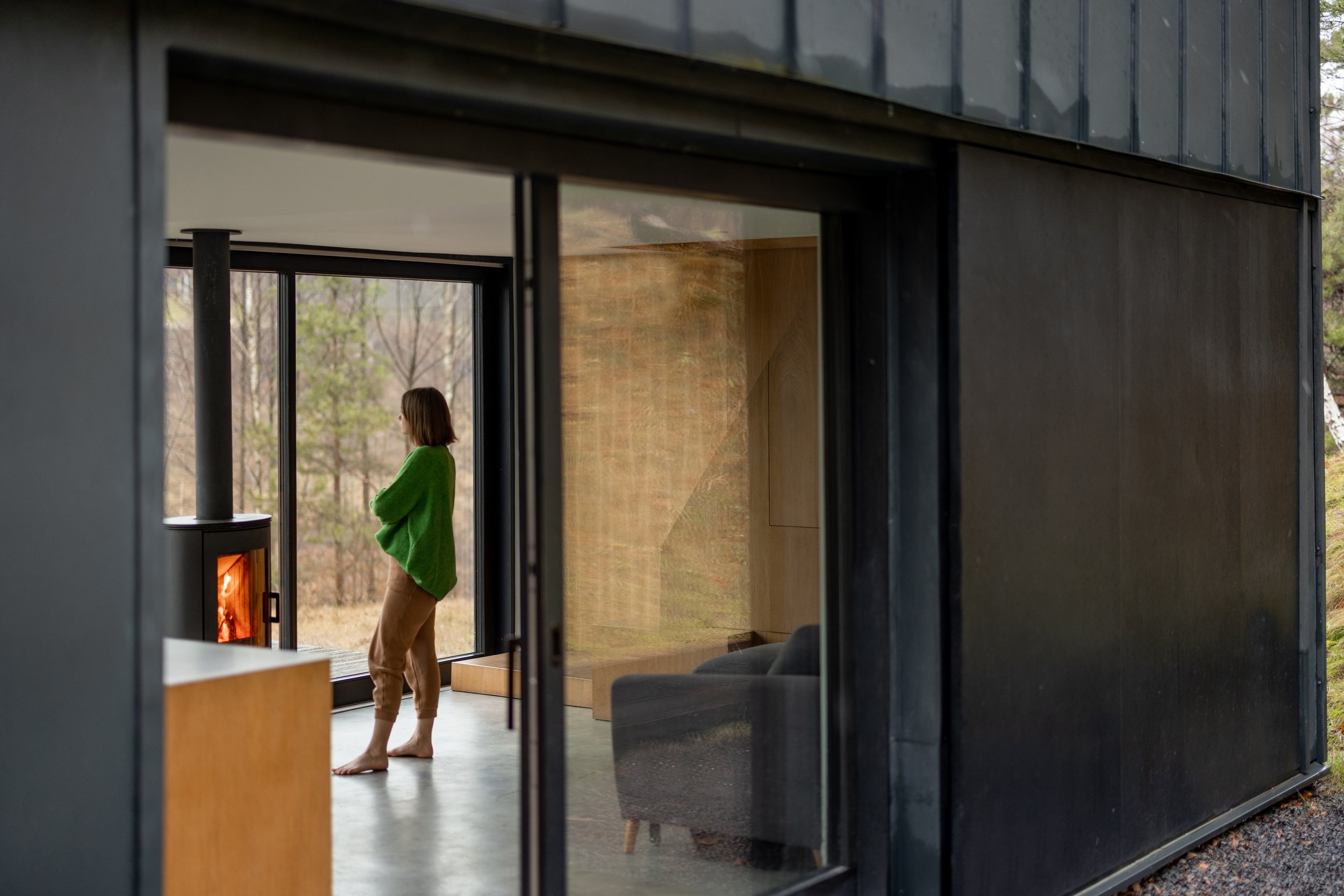 A woman in a green sweater standing inside a modern black house near a fireplace, looking outside through large glass doors.