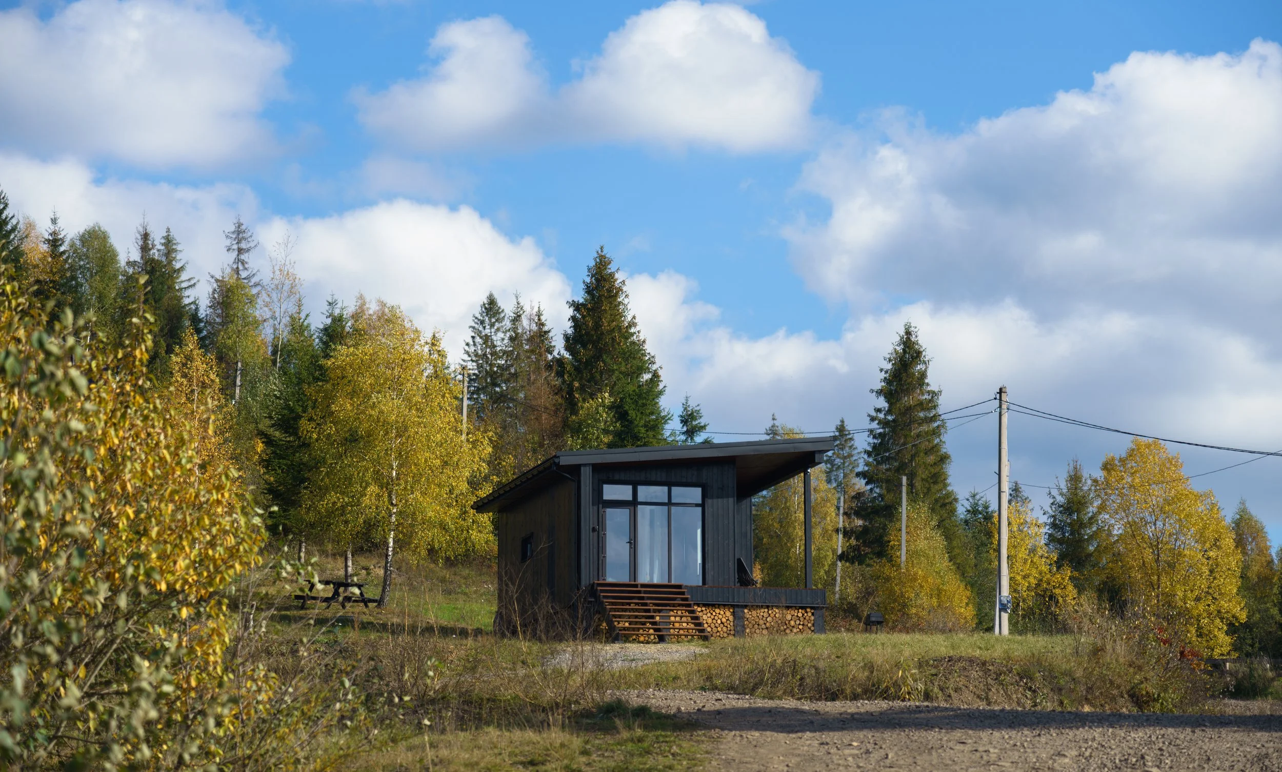 A small modern dark wooden house with large glass windows and an overhanging roof, surrounded by autumn trees with yellow and green leaves, under a partly cloudy blue sky.