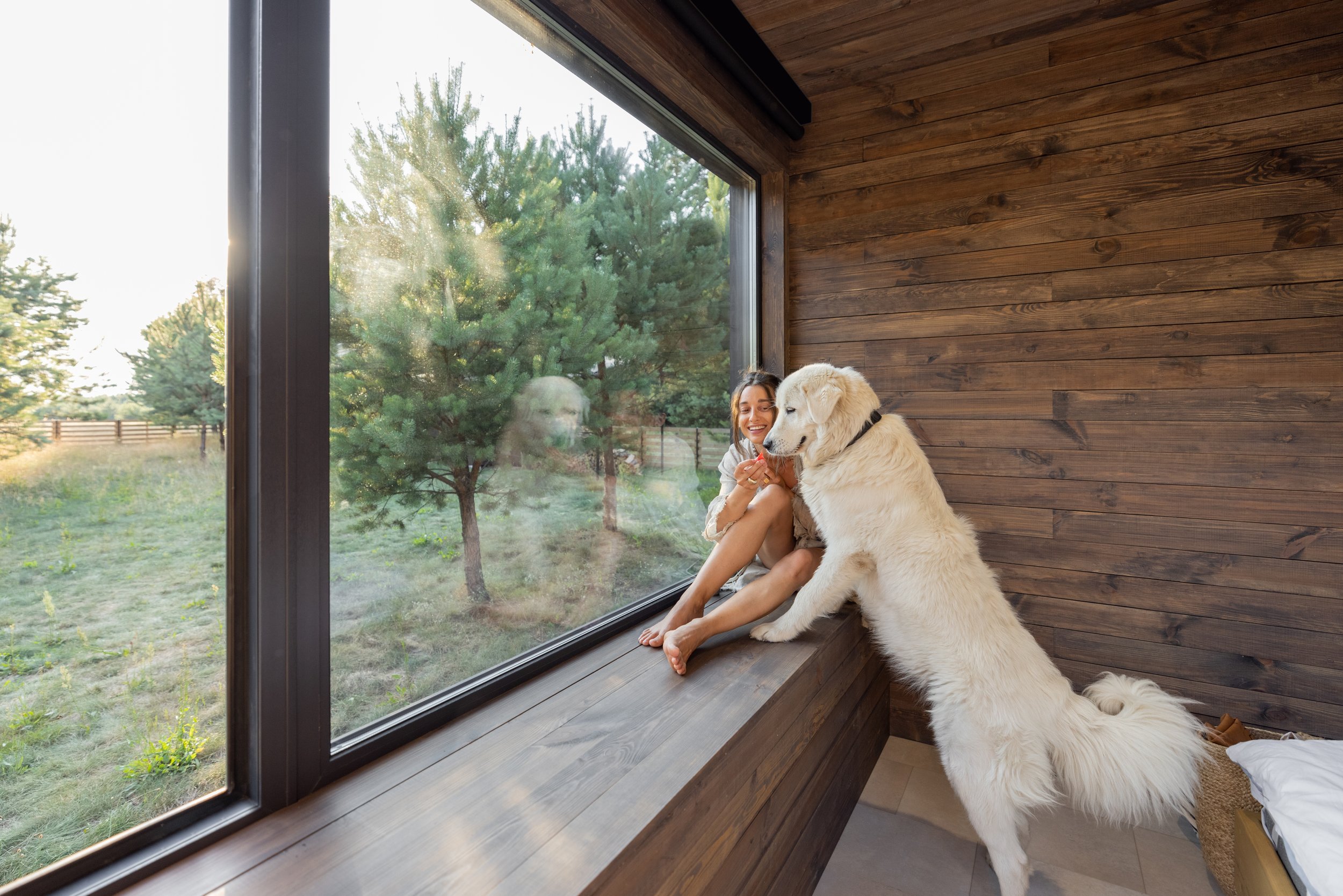A woman and a large white dog sitting on a wooden ledge inside a room with wooden paneled walls, looking out a large window at trees outside.