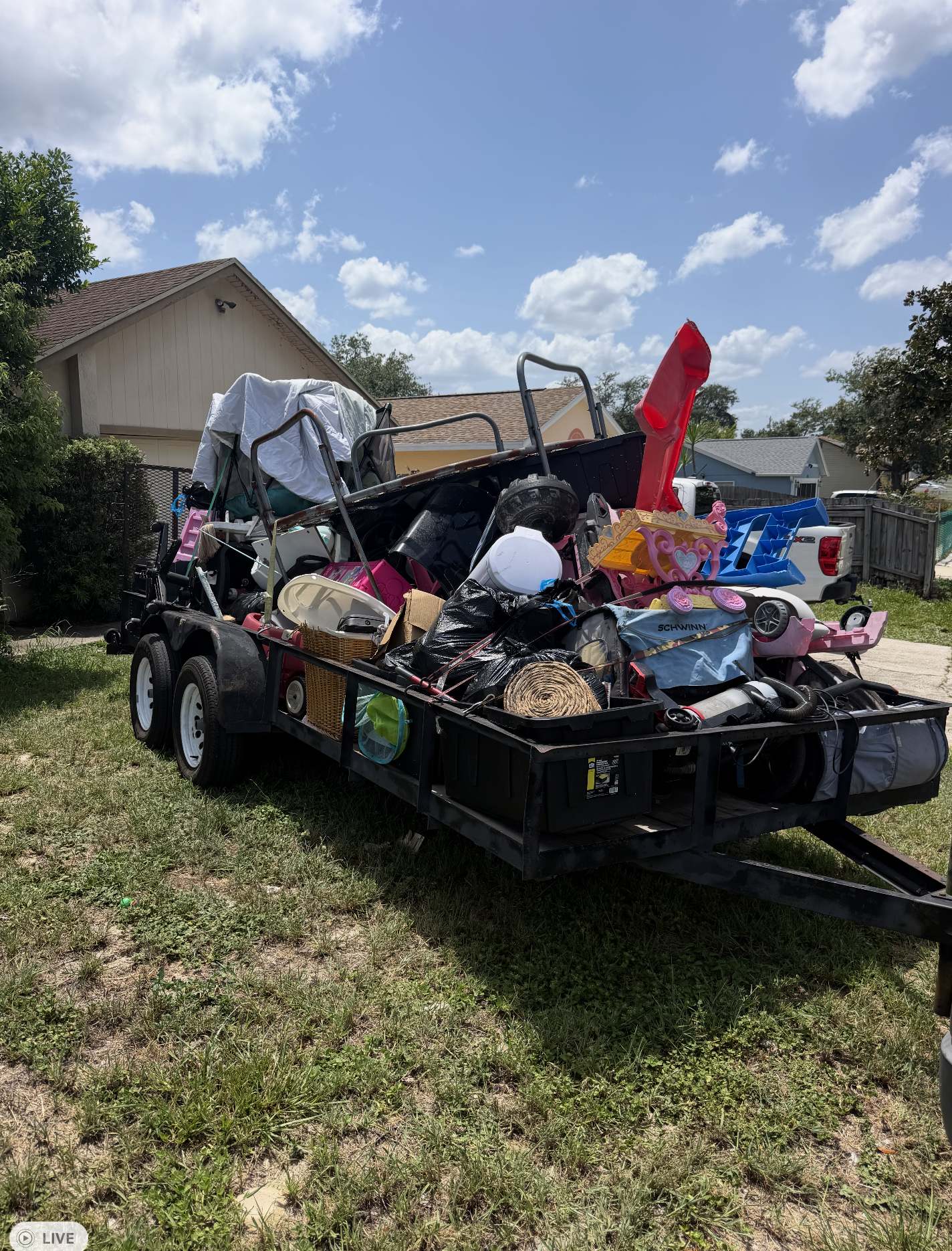 A cargo trailer filled with various items, including toys, bags, and household objects, parked on a grassy yard with houses and trees in the background under a partly cloudy sky.