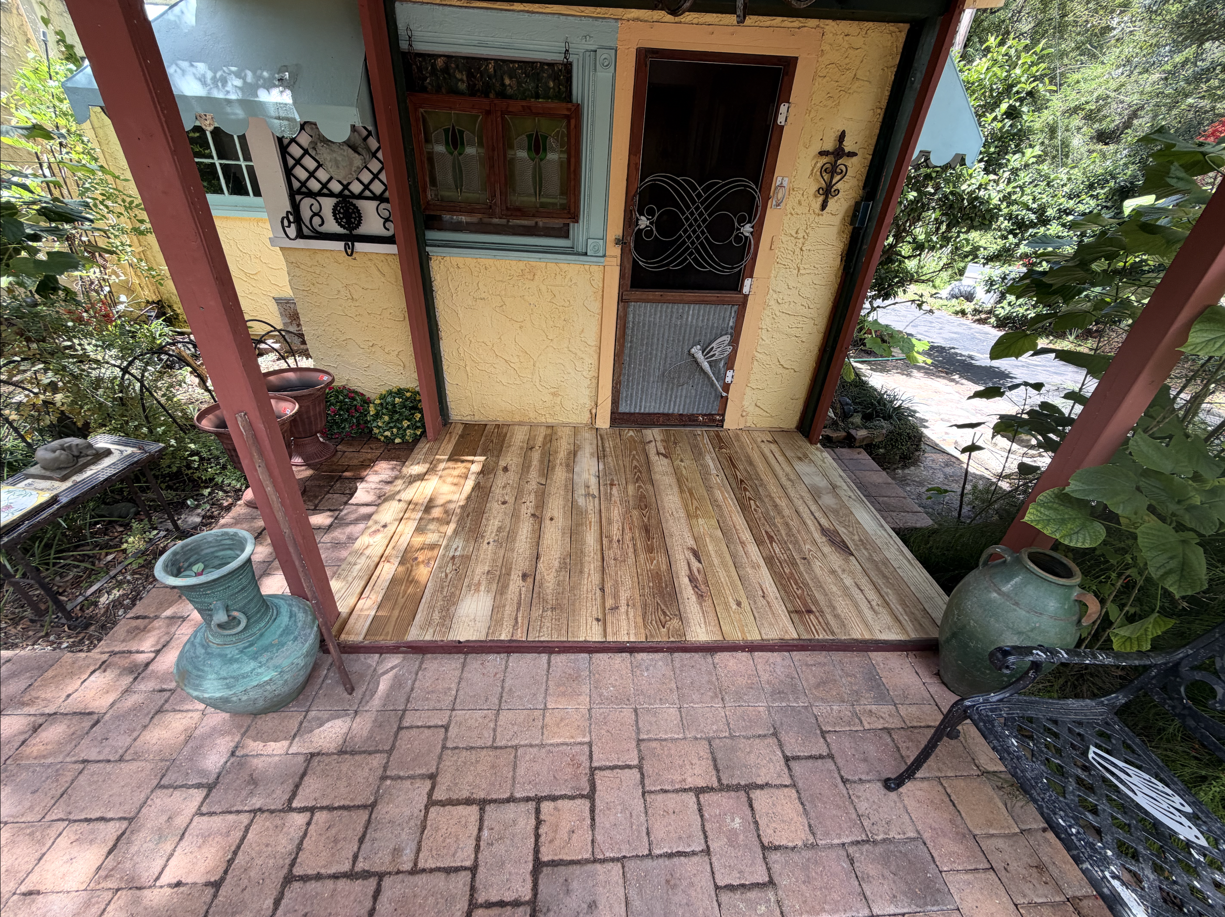 A small front porch with a wooden floor, yellow stucco walls, and a door with a metal security screen. There are potted plants on the brick walkway and greenery around the porch, with trees and bushes nearby.