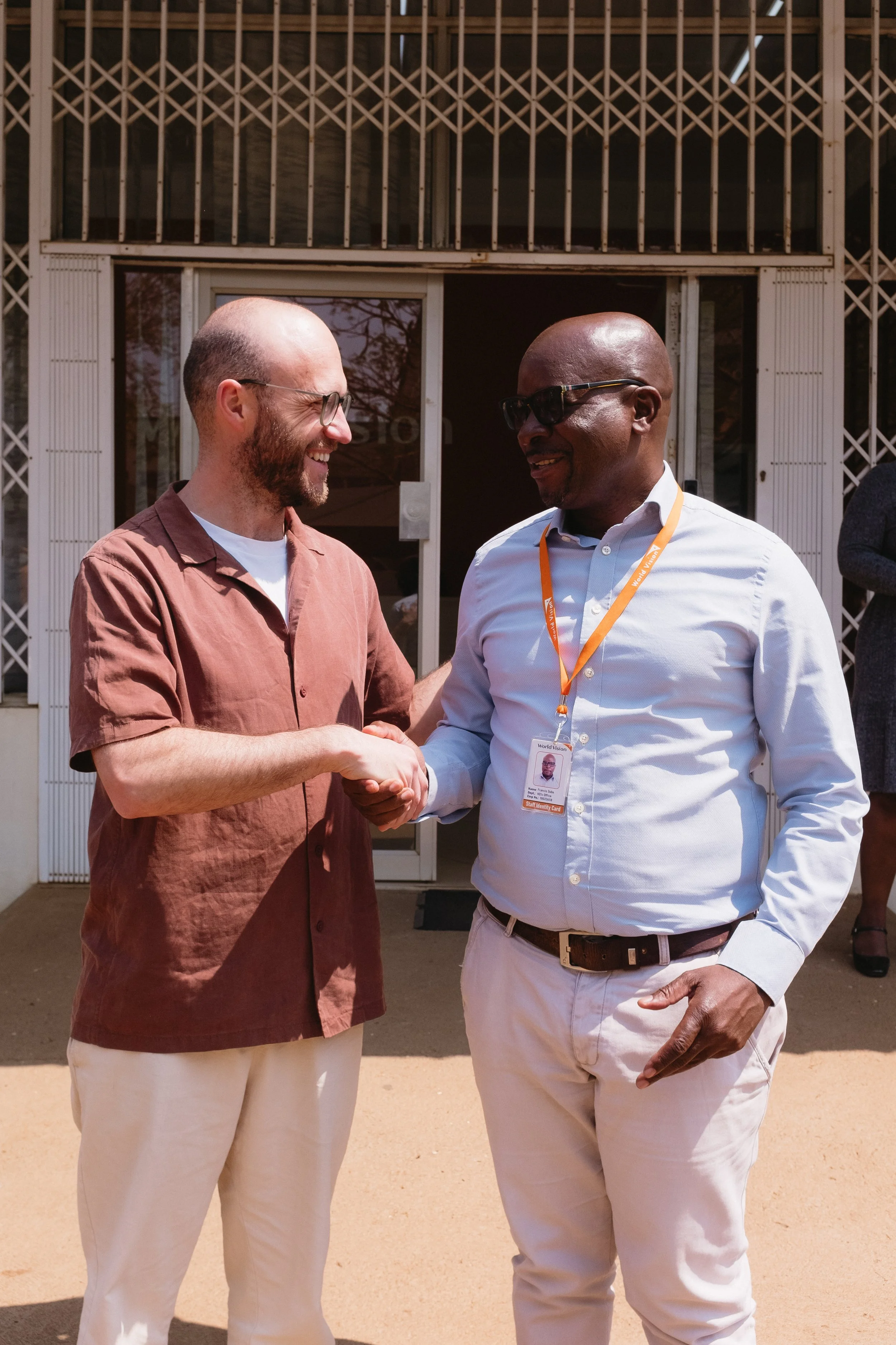 Two men shaking hands and smiling outdoors in front of a building, one with glasses and a badge around his neck, the other with a beard and glasses.
