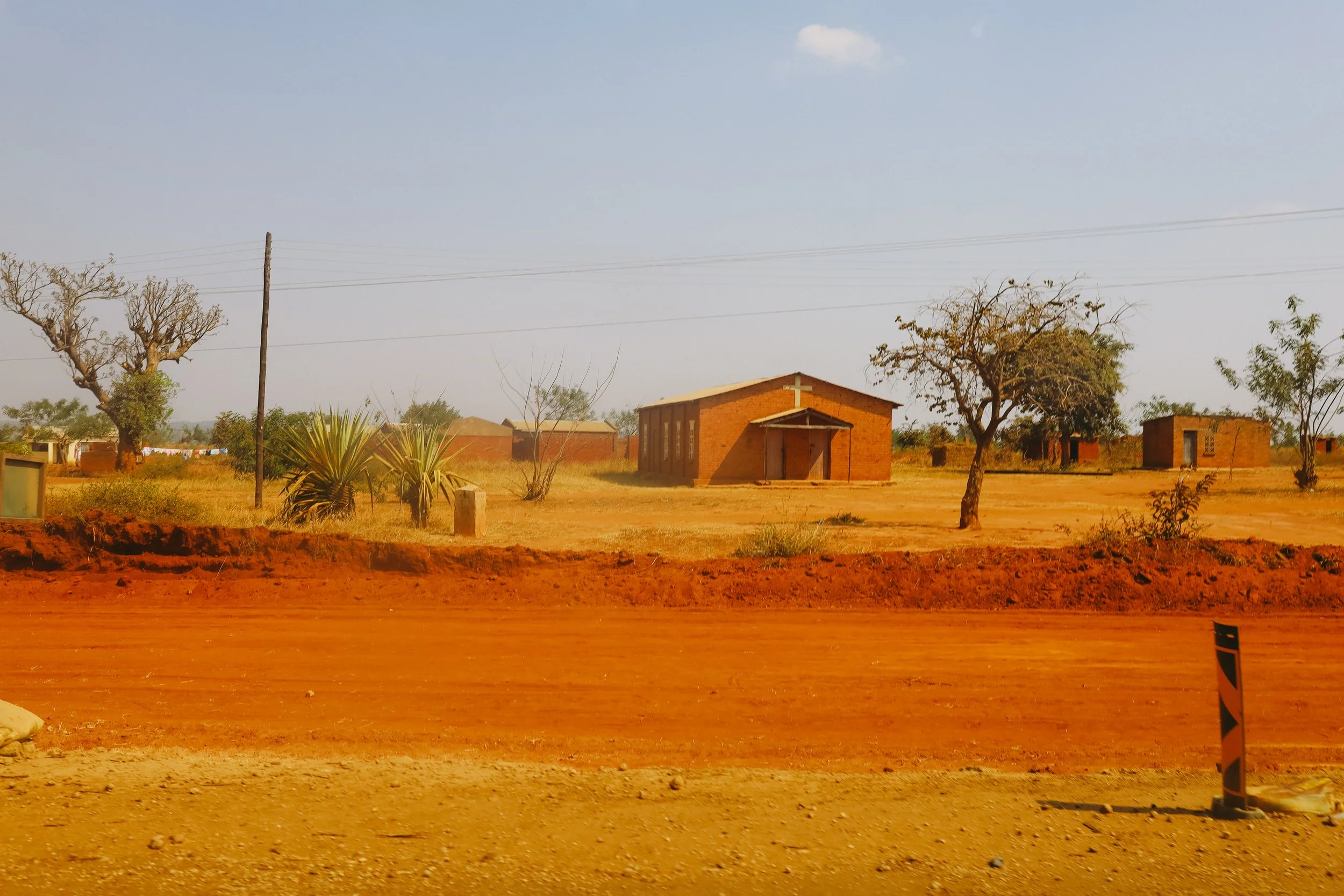 A rural landscape with red dirt, sparse trees, and small brick buildings, including a church with a cross on the roof, under a clear sky.