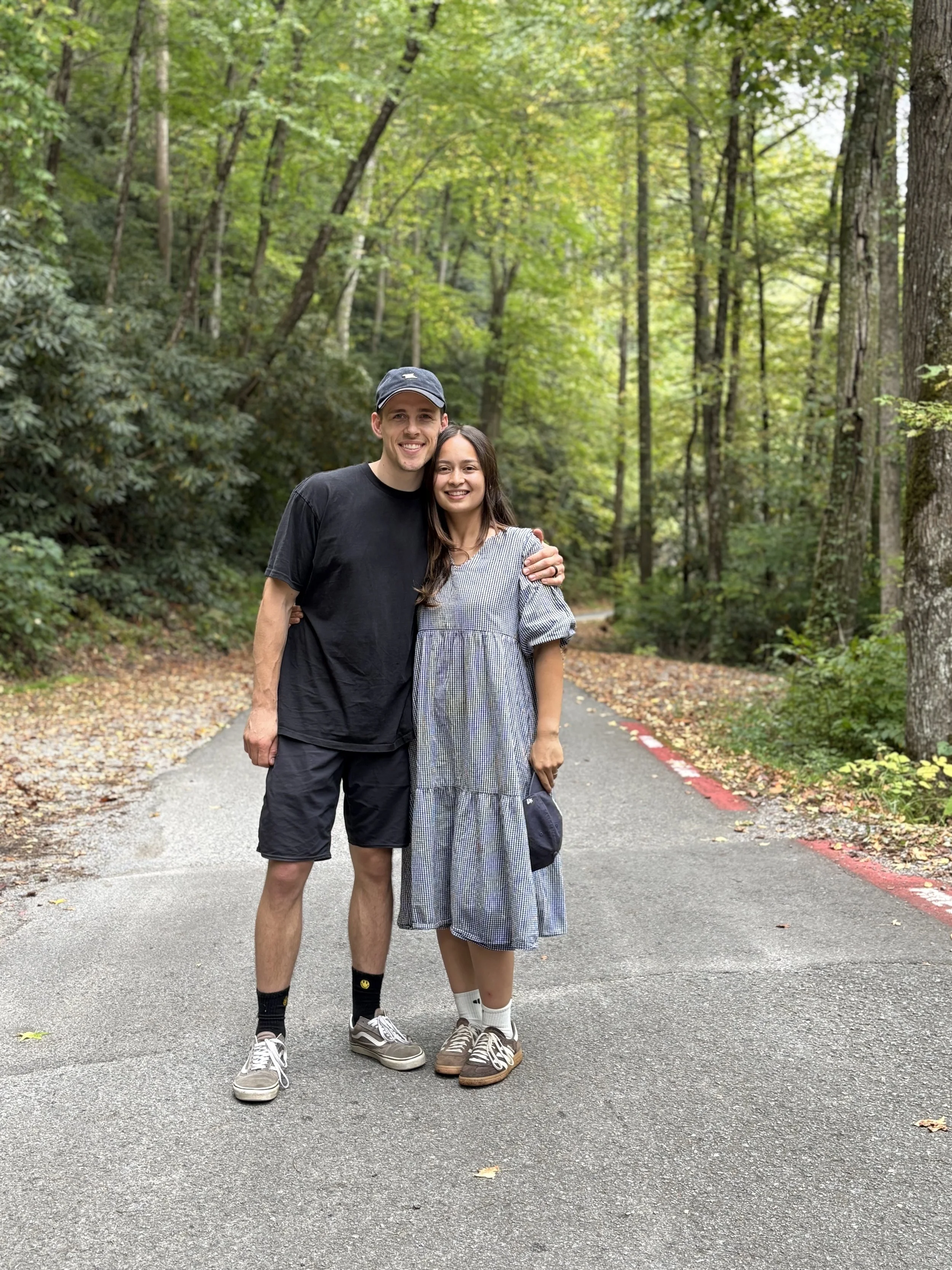 A young man and woman standing on a paved trail in a wooded area, smiling and hugging each other.