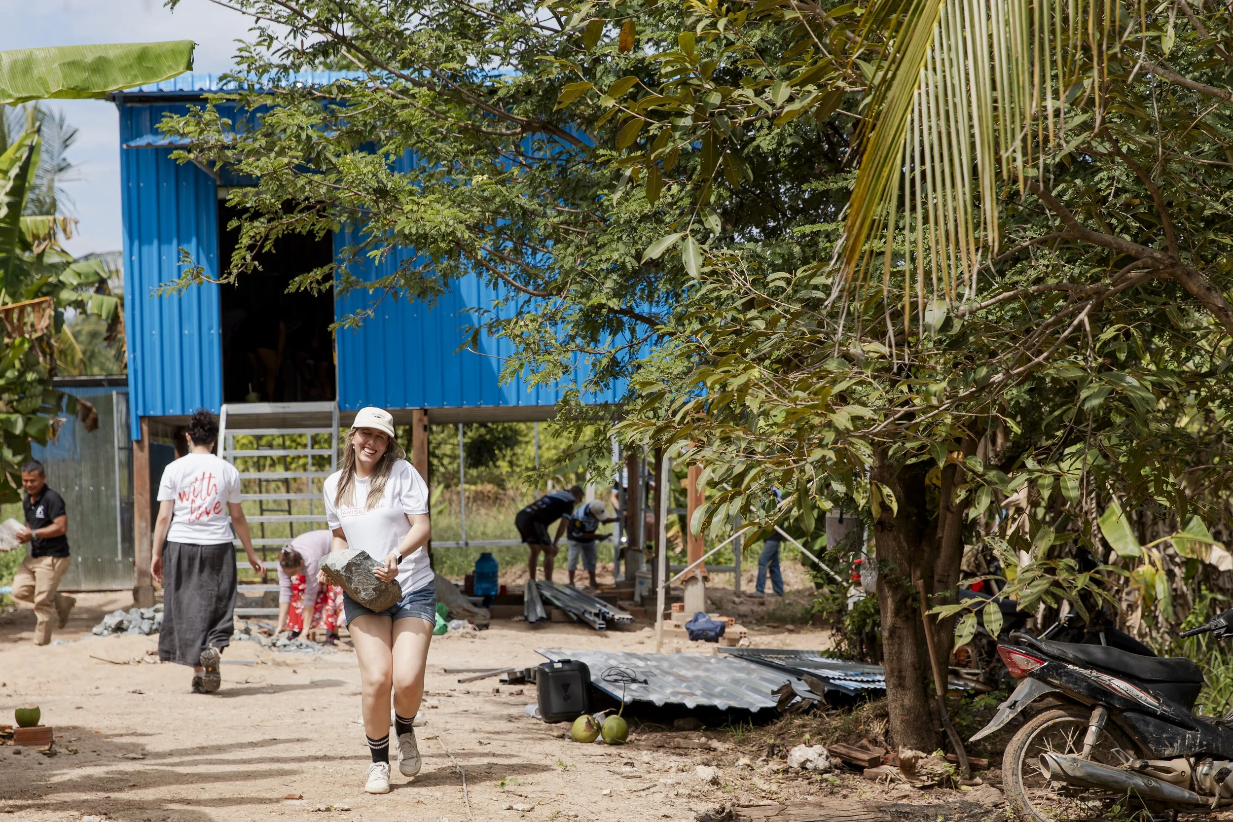 A group of people working outdoors on a construction or community project. One woman in the foreground is smiling and holding a rock, wearing a white t-shirt, shorts, and a bucket hat. Other people are in the background, some working near a partially built structure with a blue metal roof, surrounded by trees, a motorcycle, and construction materials.