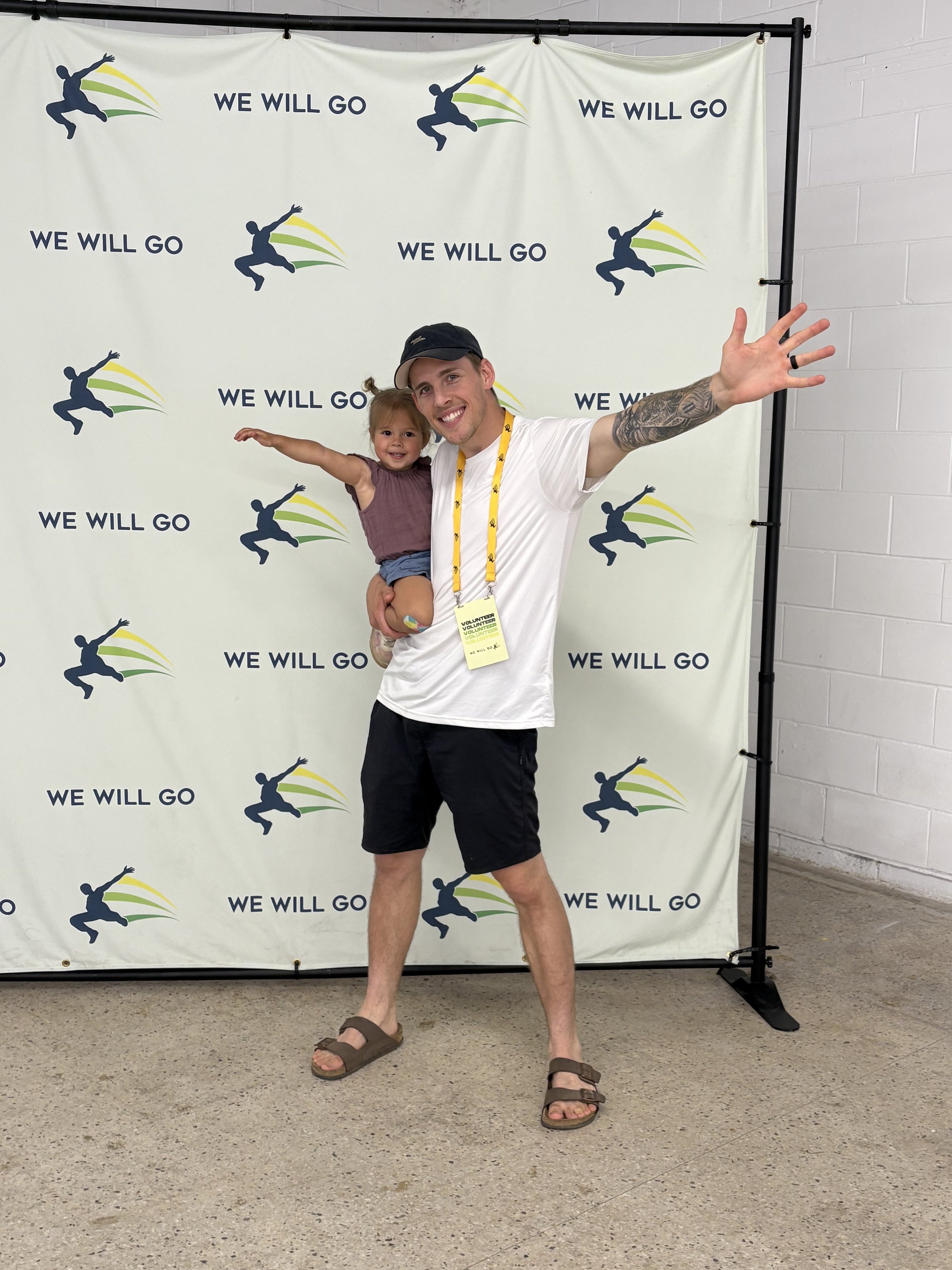 A man holding a little girl in front of a backdrop with a logo and the words "We Will Go." The man is smiling, extending his right arm outward, and wearing a white t-shirt, black shorts, brown sandals, and a black cap. The girl is wearing a sleeveless brown top, denim shorts, and has her arms outstretched.