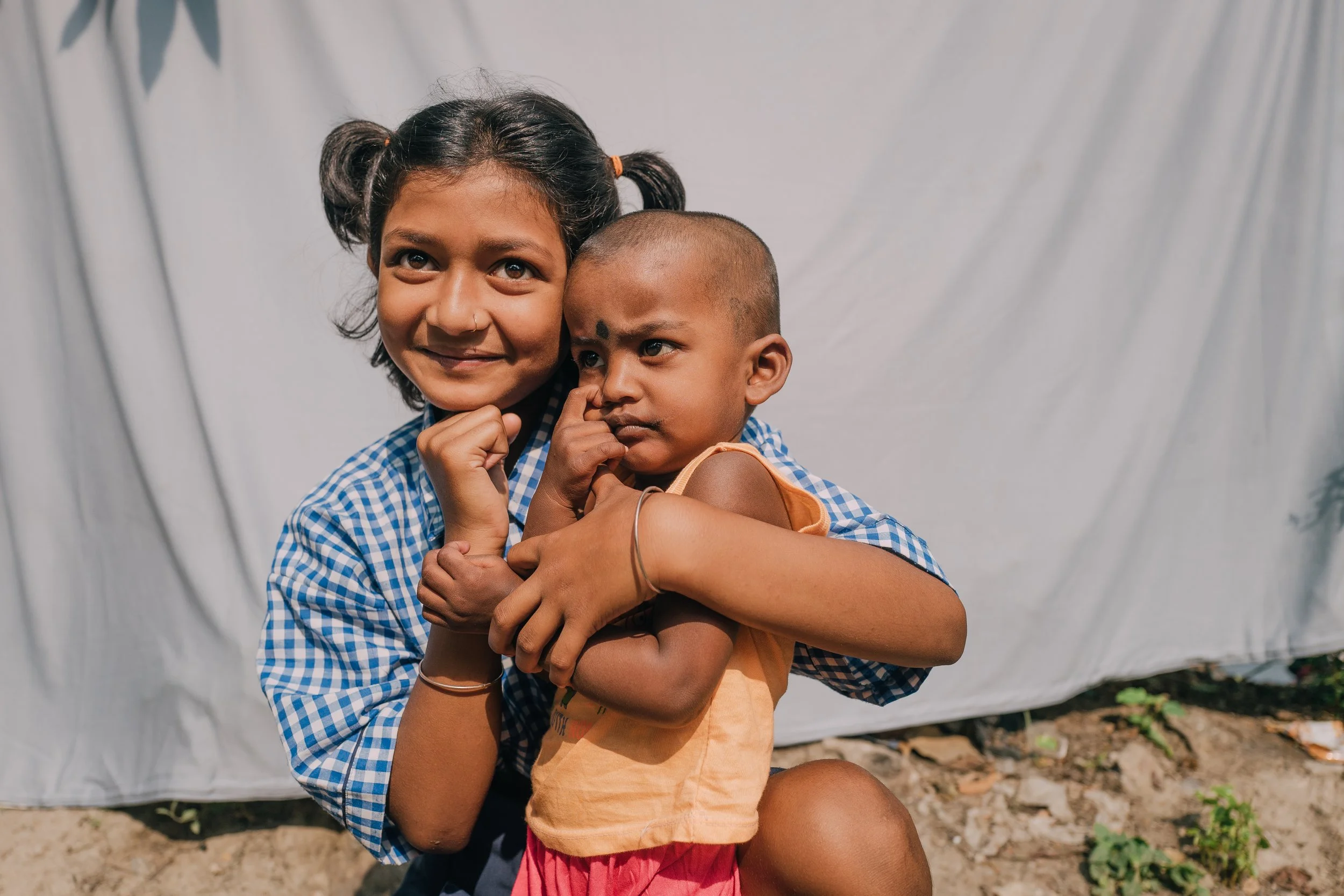 Young girl with pigtails and a girl with short hair hugging outdoors in front of a white cloth background.