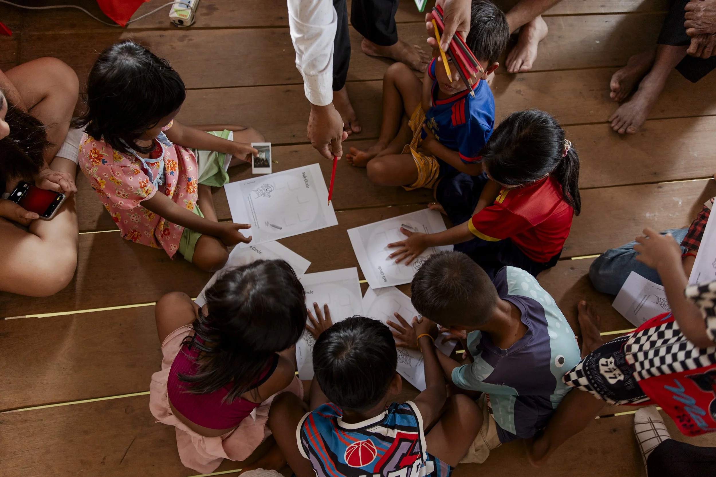 Children sitting on a wooden floor with worksheets and photos, participating in an educational activity with adult instructor.