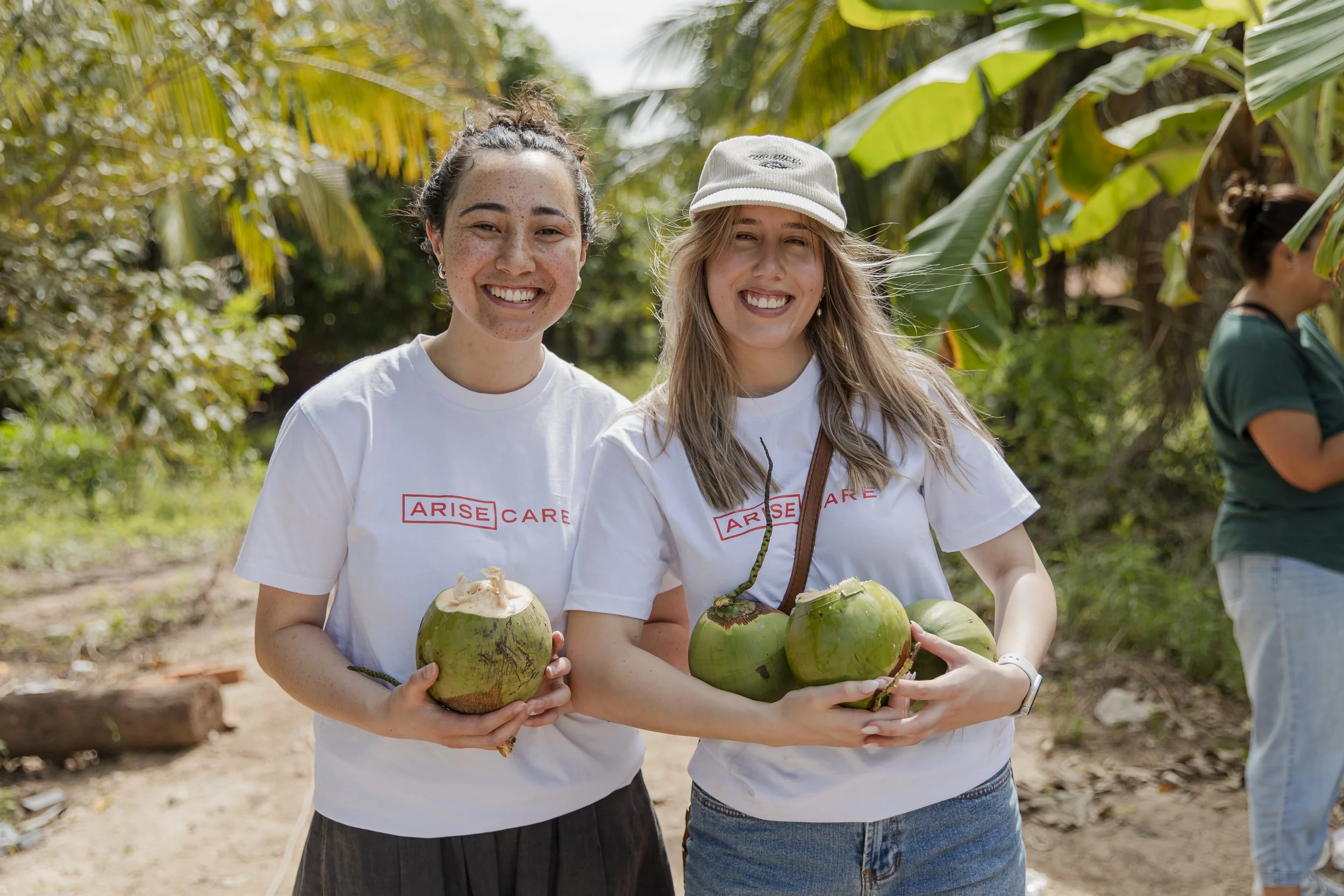 Two young women smiling and holding fresh green coconuts with straws in a tropical outdoor setting with palm trees.