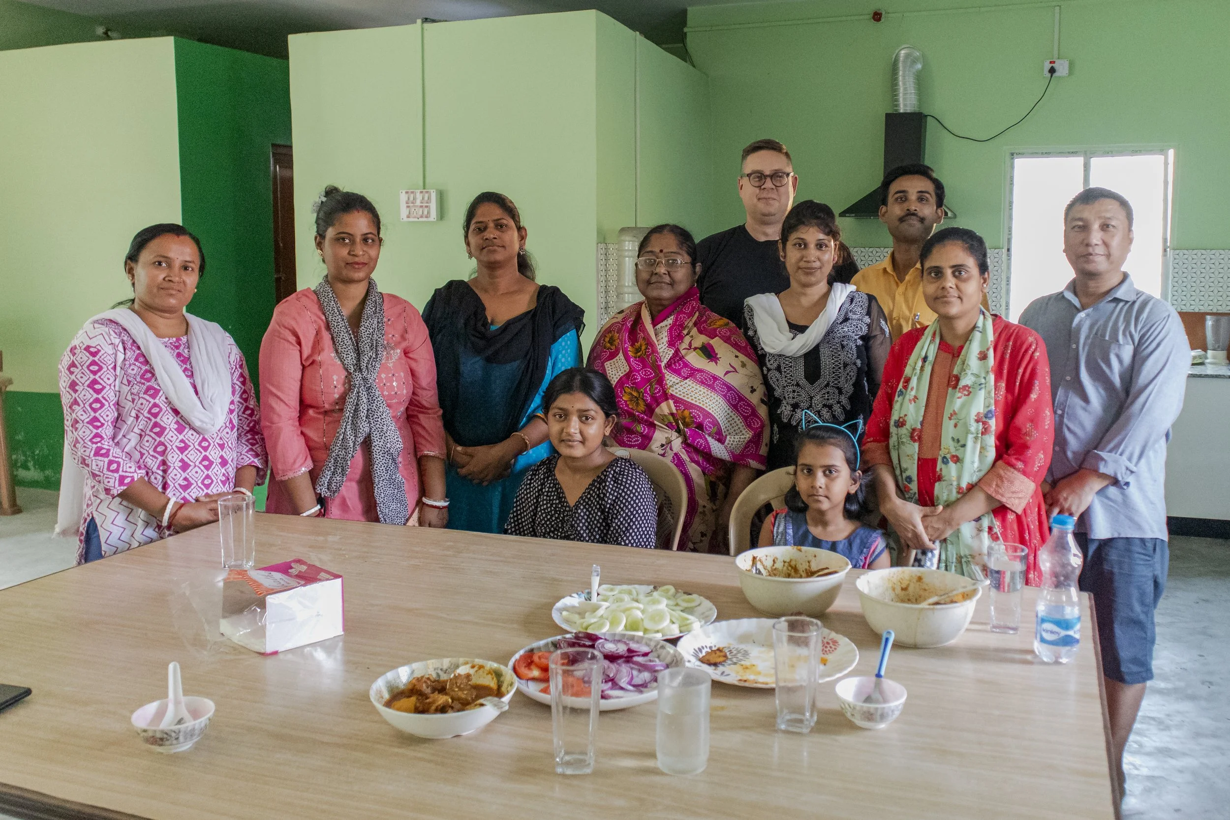 Group of people gathered around a table with food, in a room with green walls, celebrating a meal or event.