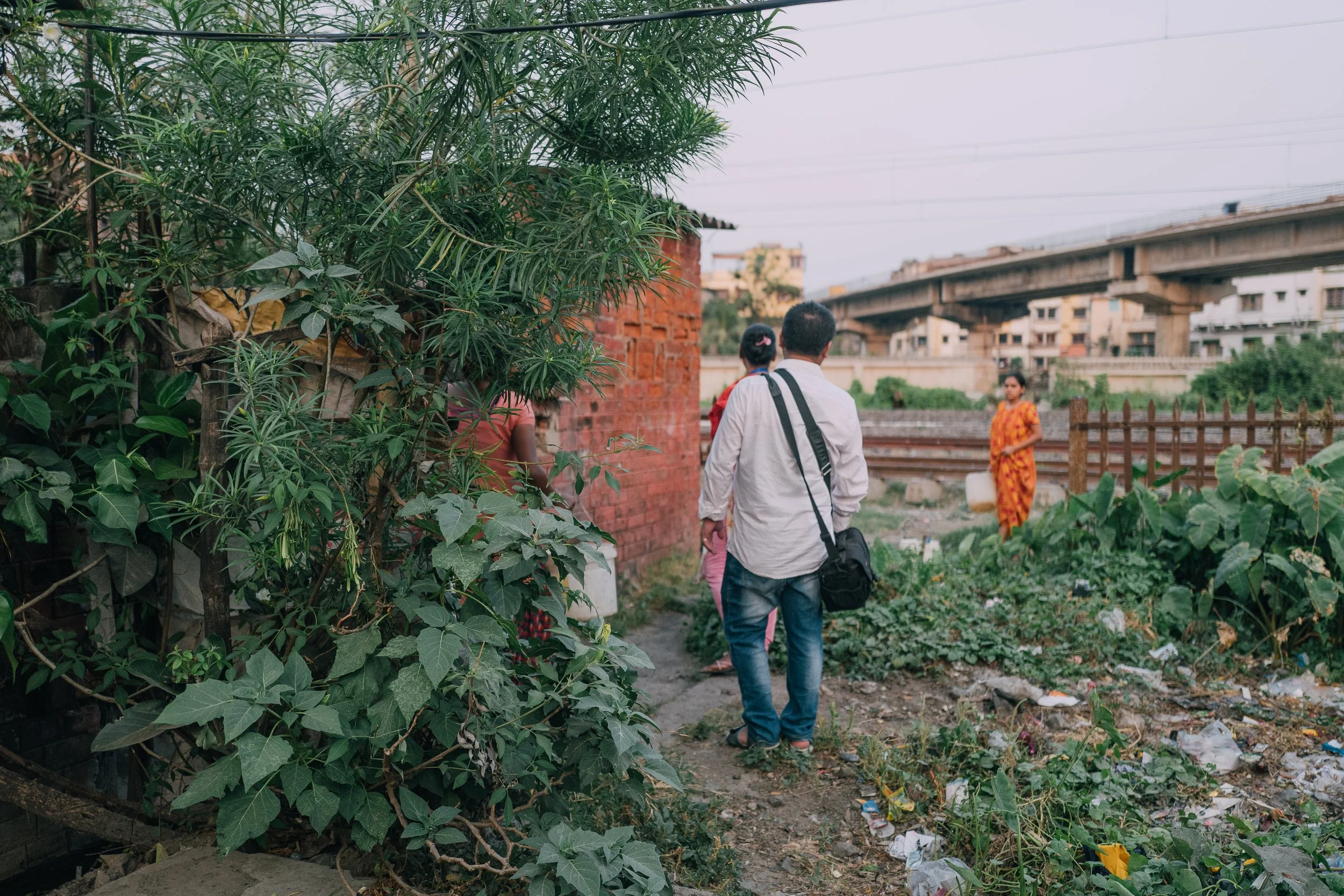 People walking along a dirt path next to green plants and a brick wall, with a woman in a yellow sari standing in the background near a wooden fence, and a railway overpass above.