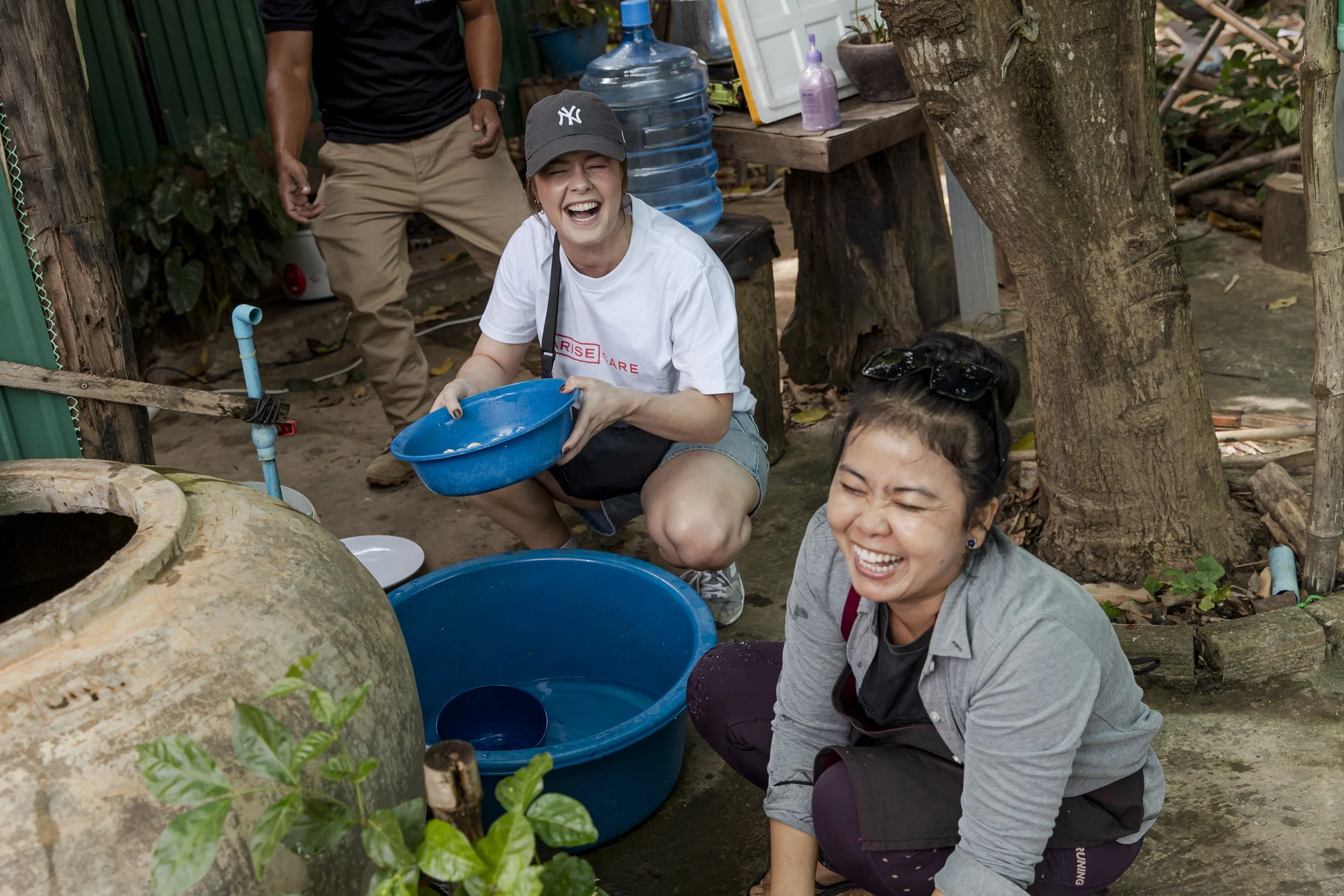 Two women laughing and smiling while washing dishes outdoors with blue basins, surrounded by plants, trees, and outdoor furniture.