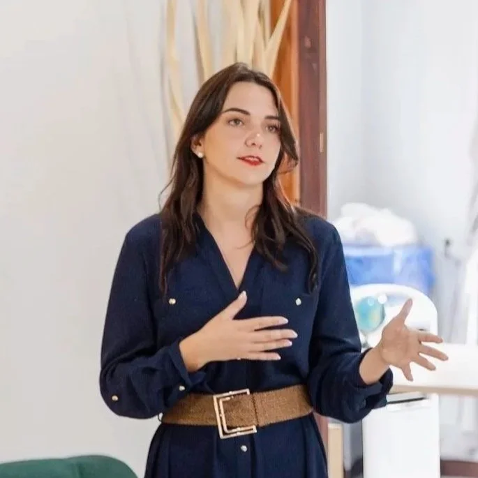 A young woman with dark hair, wearing a dark blue dress with rolled-up sleeves and a tan belt, is standing indoors with one hand on her chest and the other gesturing outward, in front of a light-colored wall.