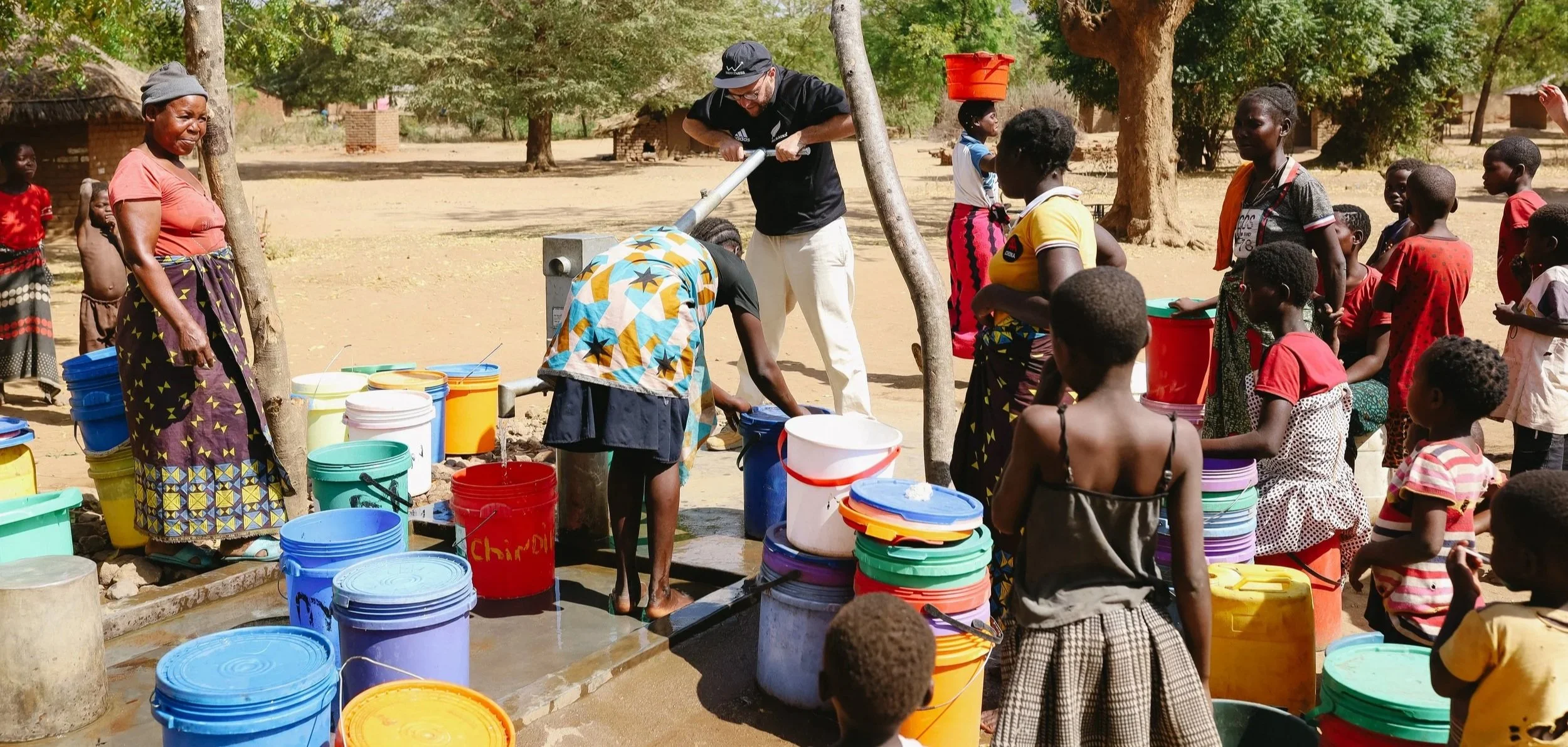 Group of children and adults gather around a water pump; children get water into buckets while adults supervise, outdoors in a rural setting with trees and simple buildings in the background.