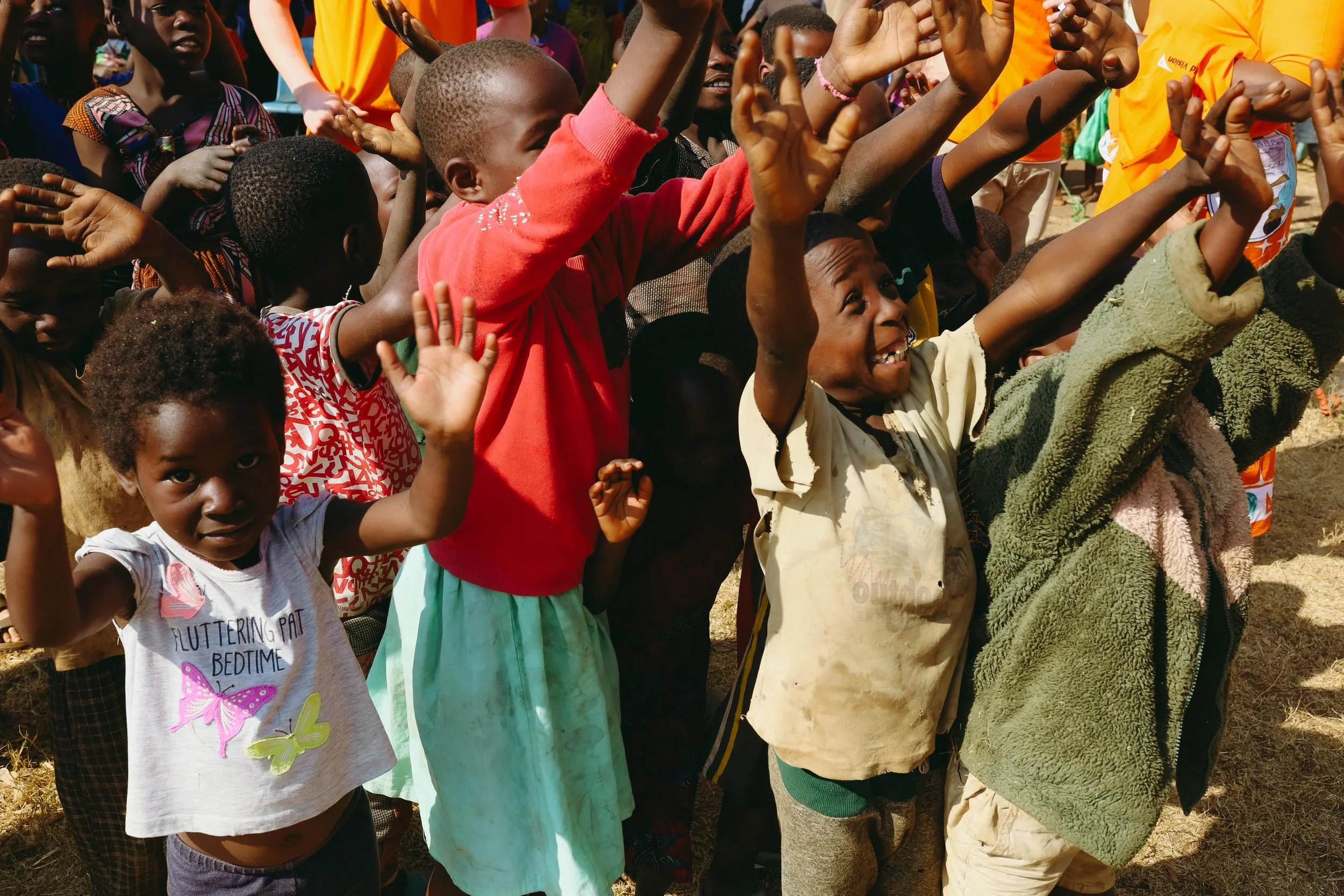 Group of children outdoors, many with arms raised and smiling, wearing colorful clothes, some showing excitement or joy.