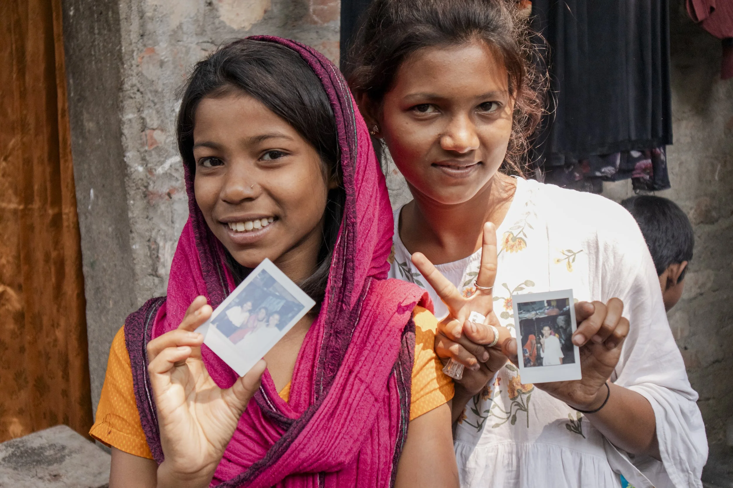 Two young girls standing close together, smiling, and holding Polaroid photos. The girl on the left is wearing a pink headscarf and orange shirt, while the girl on the right is wearing a white top and making a peace sign with her fingers. They are standing outdoors with a stone wall in the background.