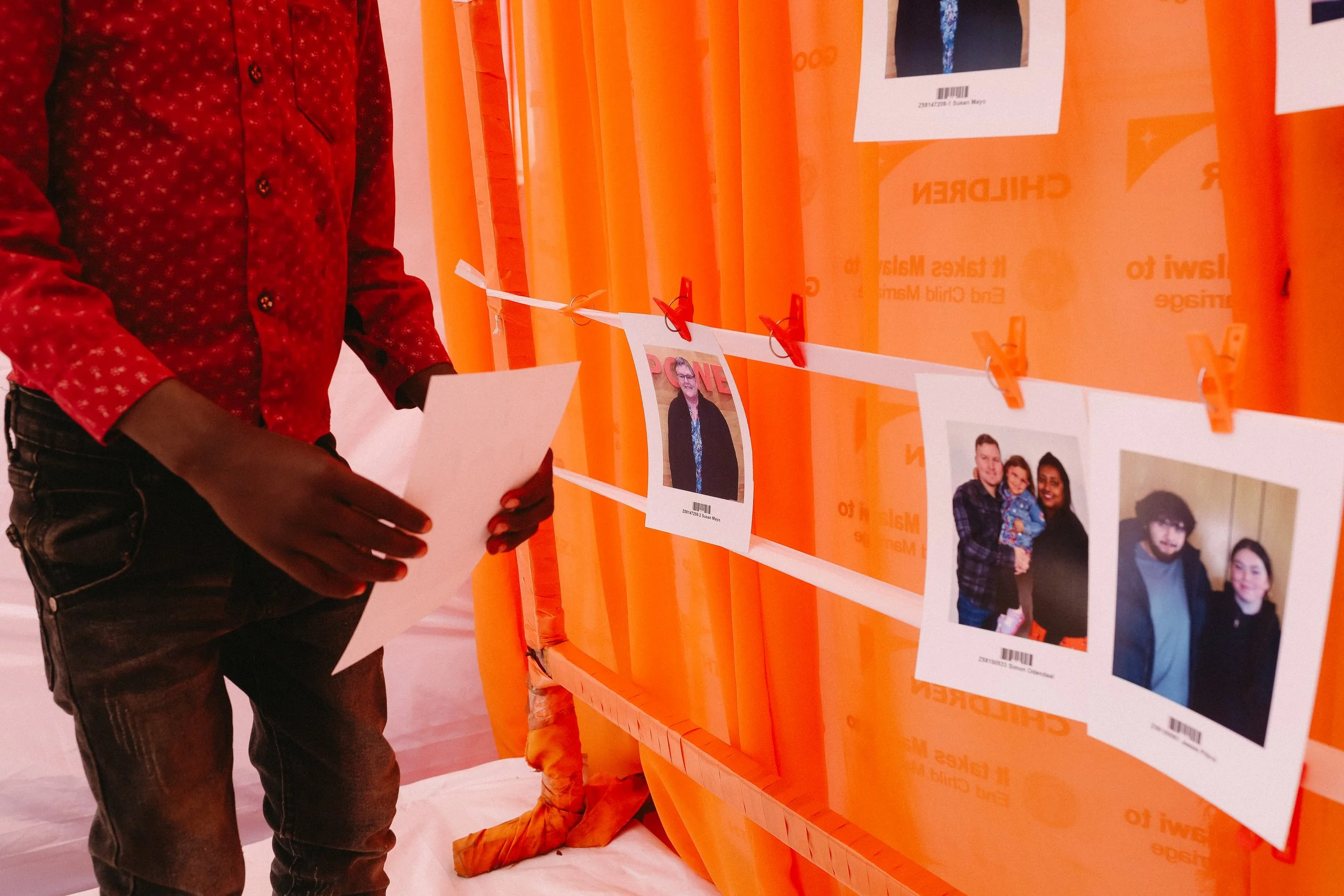 A person in a red patterned shirt holding a piece of paper, standing near a display of photographs hanging on a white string with orange clips. The photographs feature various people and are set against an orange backdrop.