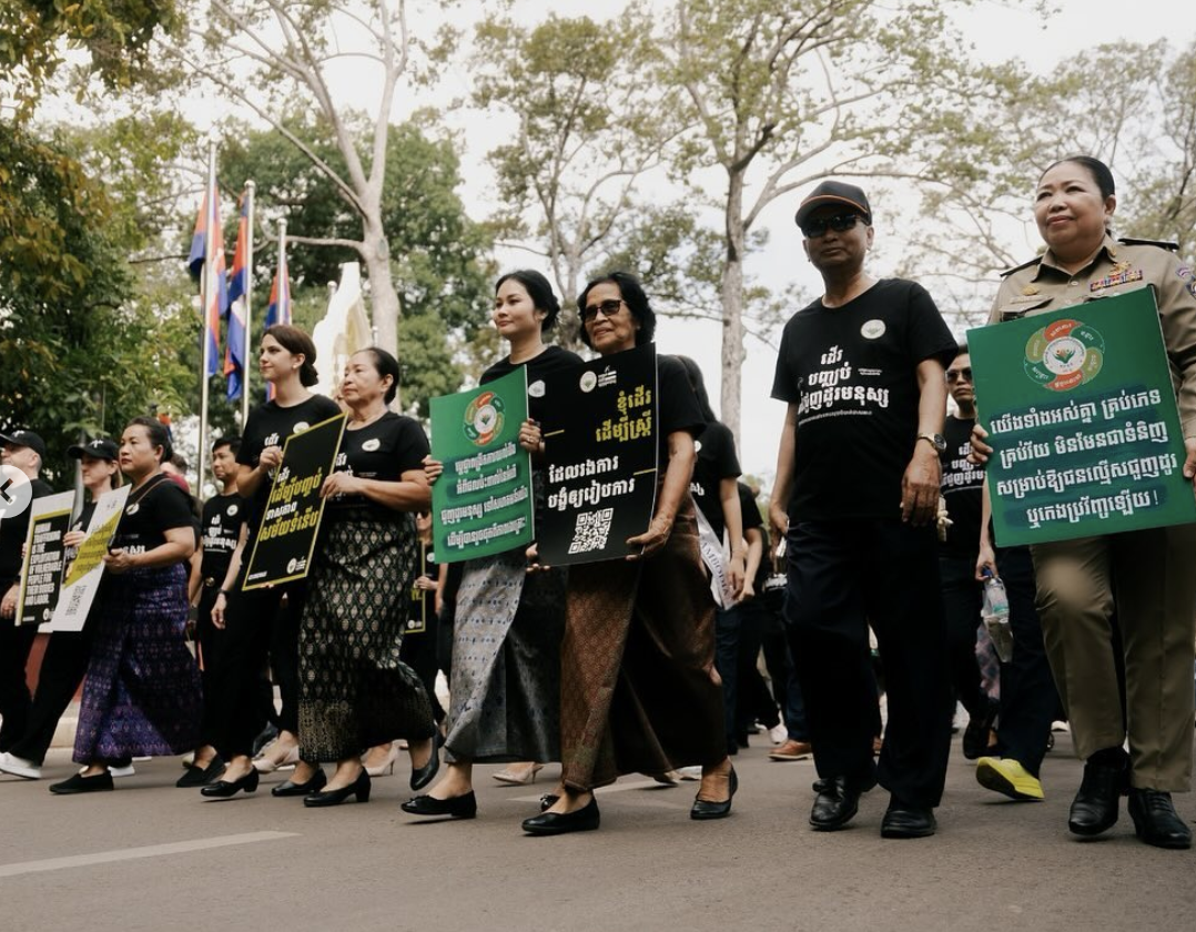 People participating in a march or demonstration, holding signs with text, walking outdoors with trees and flags in the background.