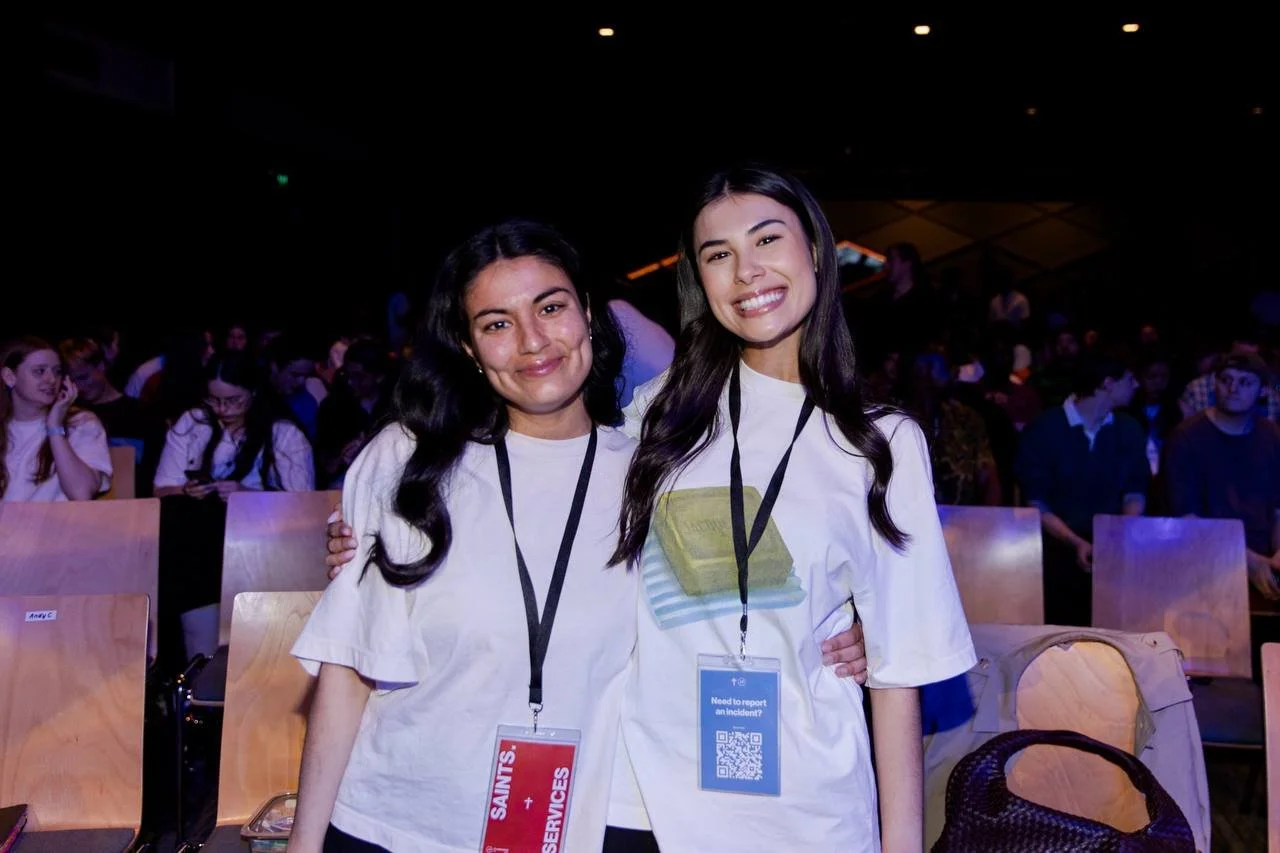 Two young women smiling and posing together at an event, wearing white shirts and conference badges, with a crowd sitting in the background.
