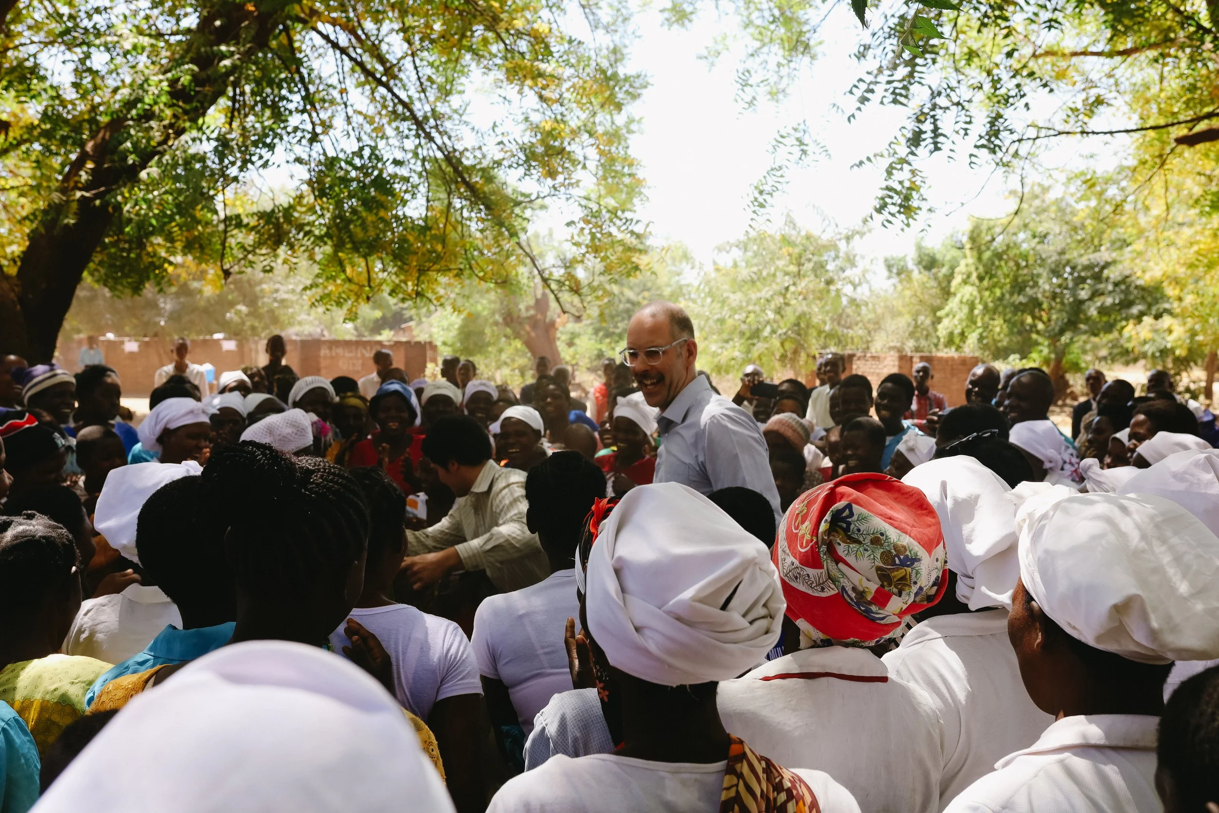 A man with glasses and a white shirt smiling among a large crowd of people wearing white headscarves and traditional clothing outdoors under trees.