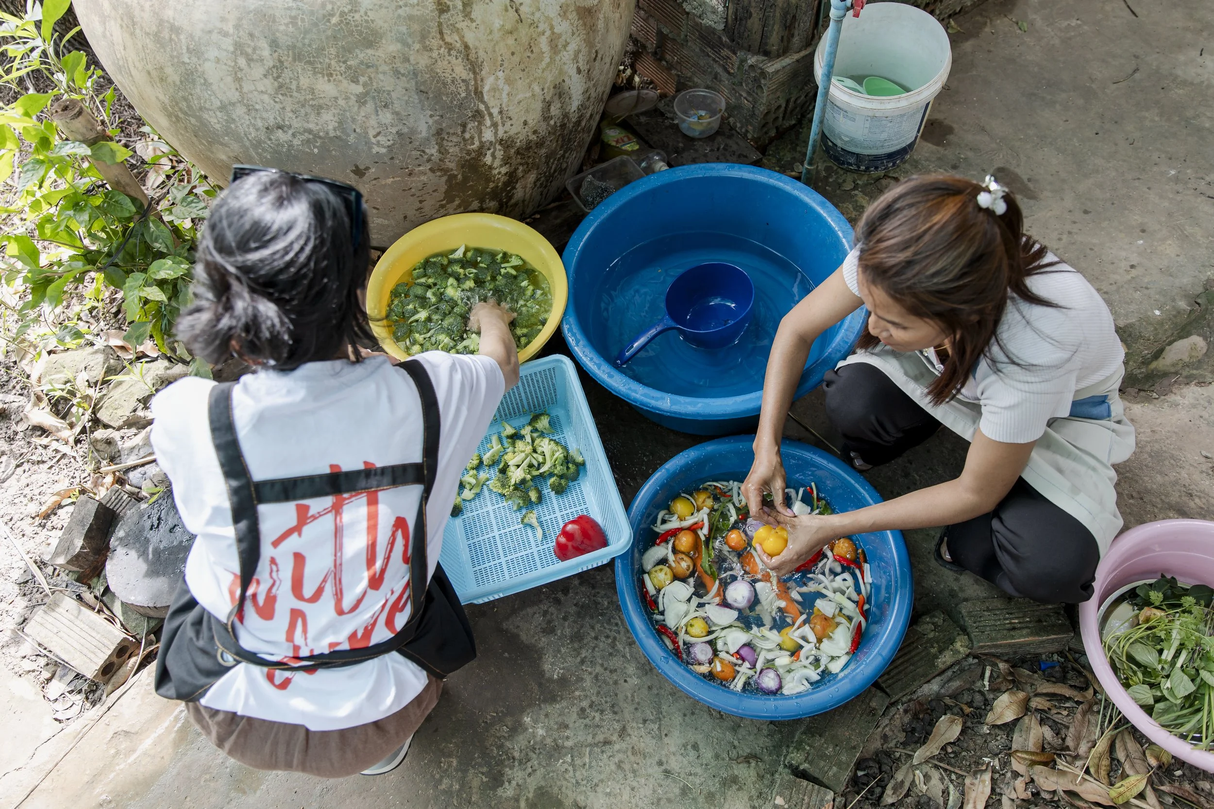 Two women washing vegetables in large colorful basins outdoors.
