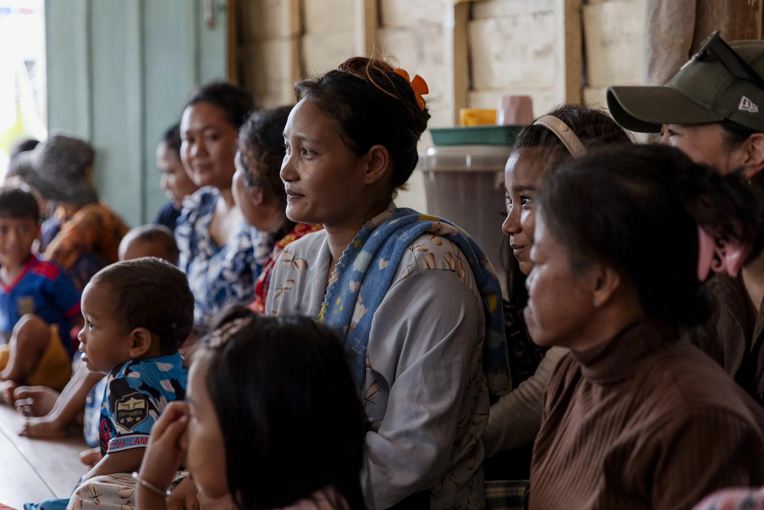 Group of women and children sitting indoors, attentively listening, with some traditional and casual clothing visible, in a rustic setting.