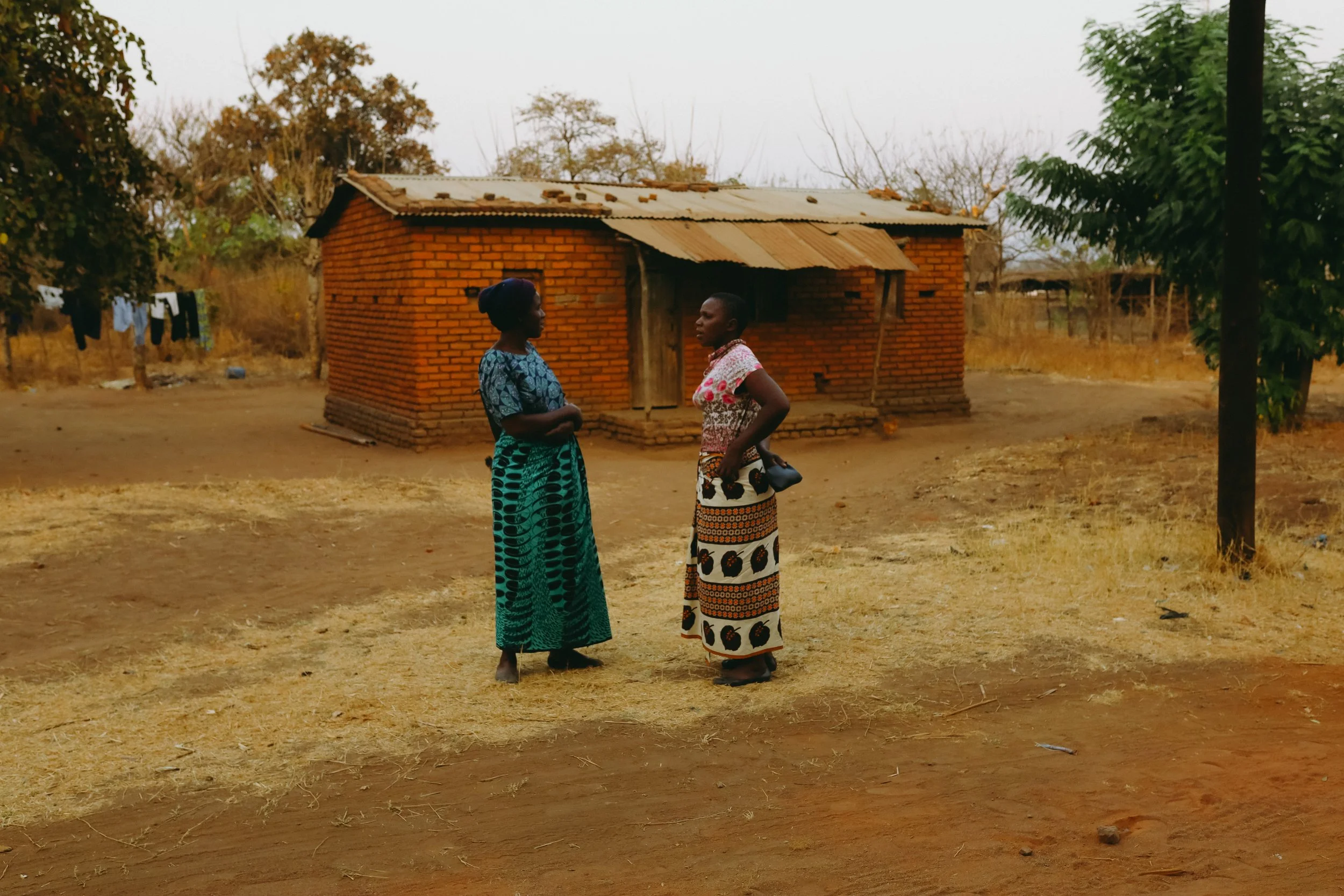 Two women standing and talking outdoors in front of a small brick house with a corrugated metal roof. One woman wears a blue patterned top and skirt, and the other wears a pink top and a patterned skirt. There is a tree on the right and laundry hanging on a line in the background.