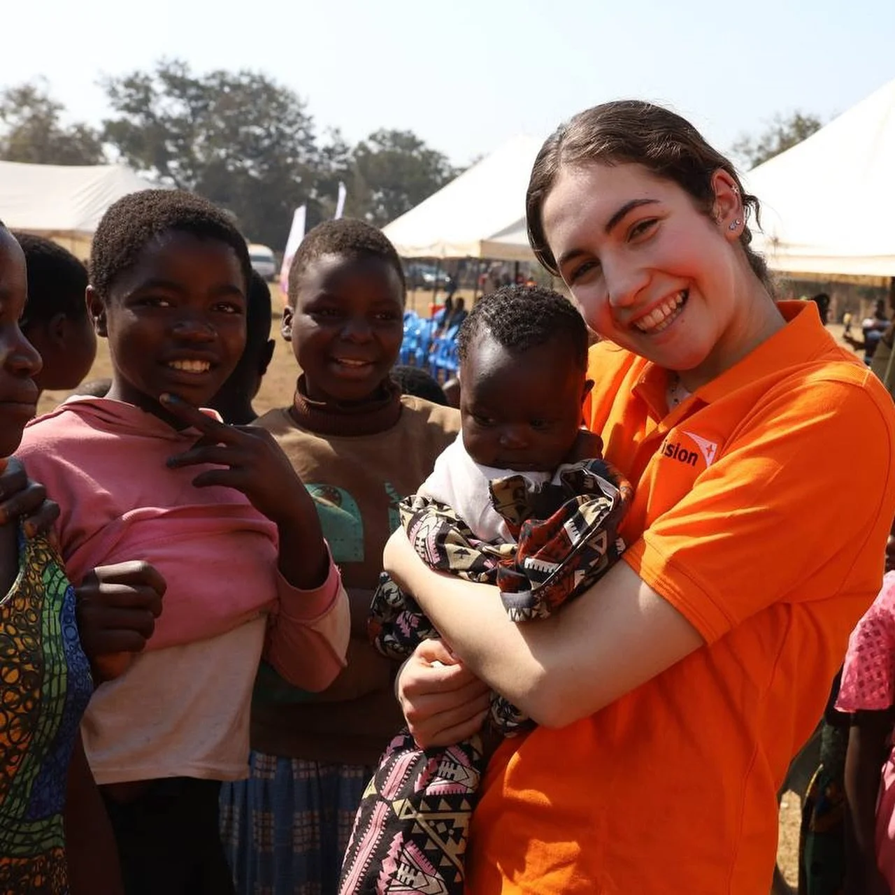A group of children and a woman with a baby outdoors at a community event, smiling and interacting.
