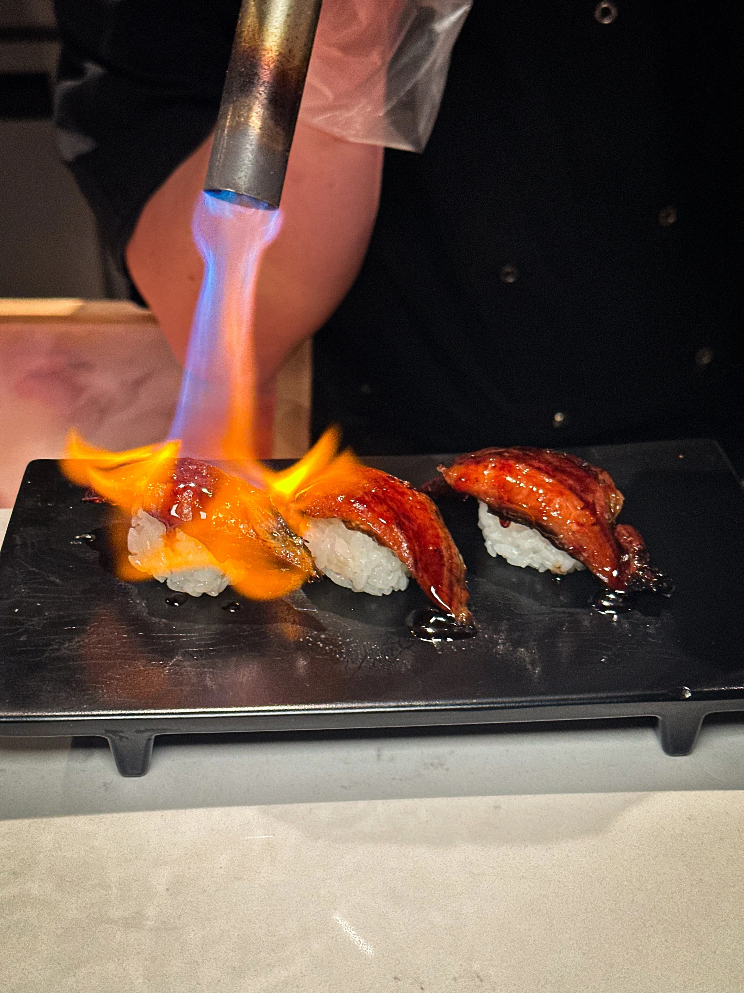 Chef uses a blowtorch to sear sushi with eel and sauce on a black plate.