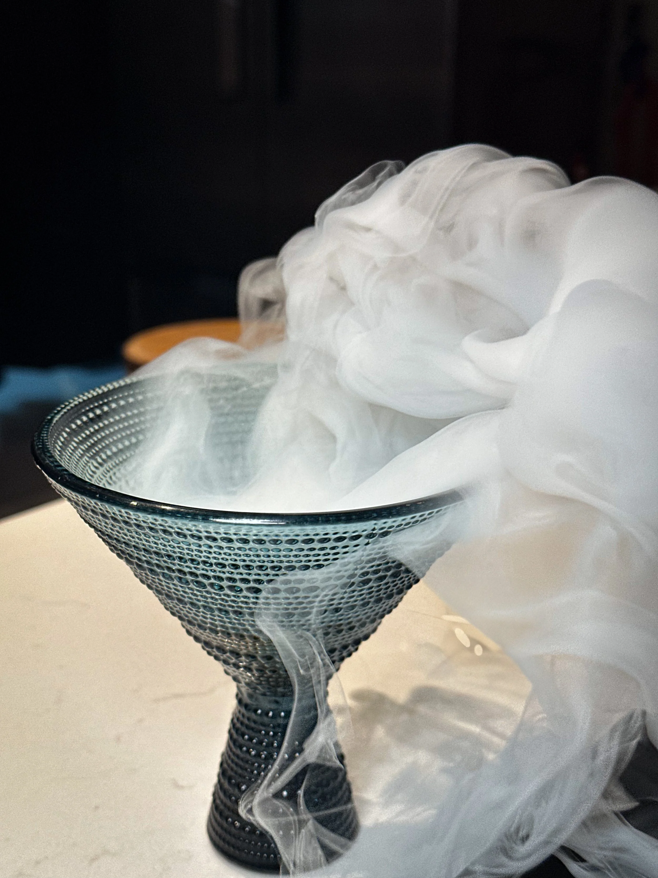 A metal cone-shaped strainer or bowl filled with white smoke or vapor, likely dry ice or vapor, spilling over the sides.