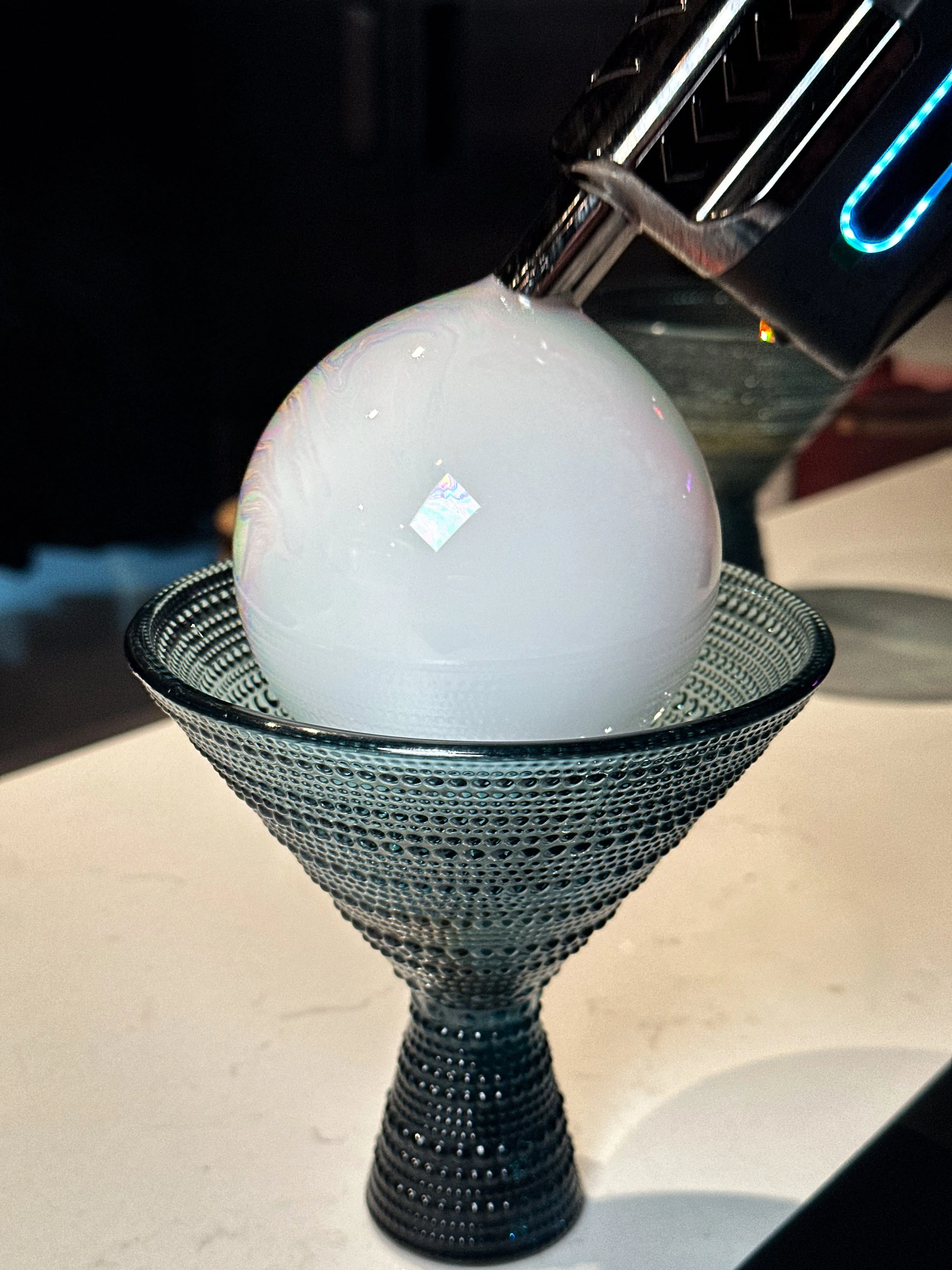 A large, opaque white bubble resting in a black textured cone-shaped bowl, with a metallic soda fountain attached at the top.