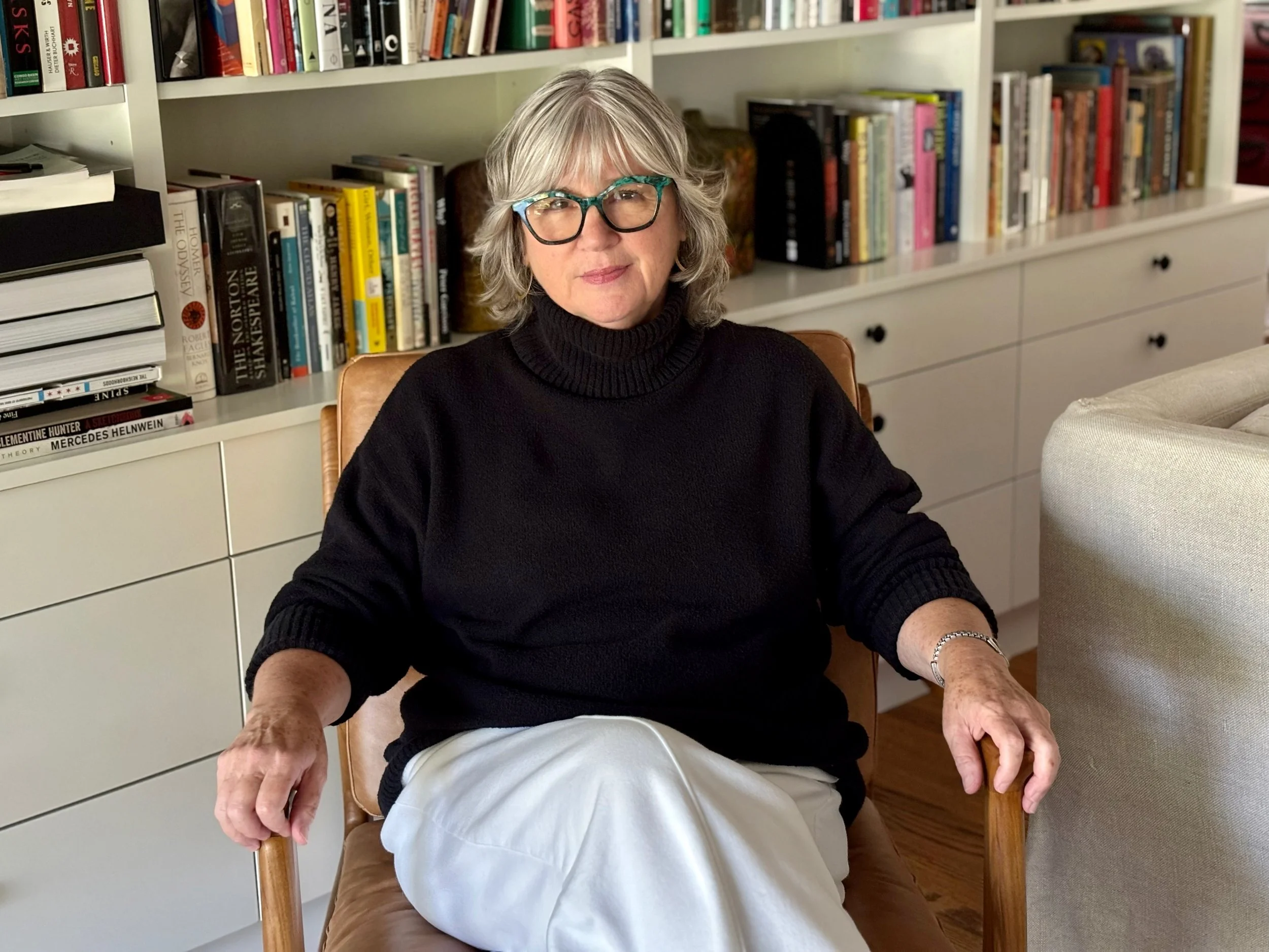 A woman with gray hair and glasses sitting in a wooden chair in a room with a bookshelf in the background. She is wearing a black turtleneck sweater and white pants.