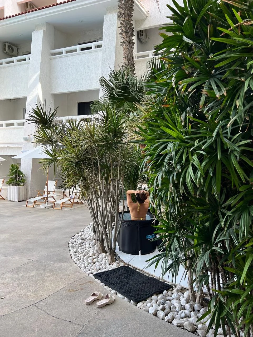 A woman relaxing in a black hot tub surrounded by tropical plants on a hotel balcony or patio with white walls and lounge chairs nearby.