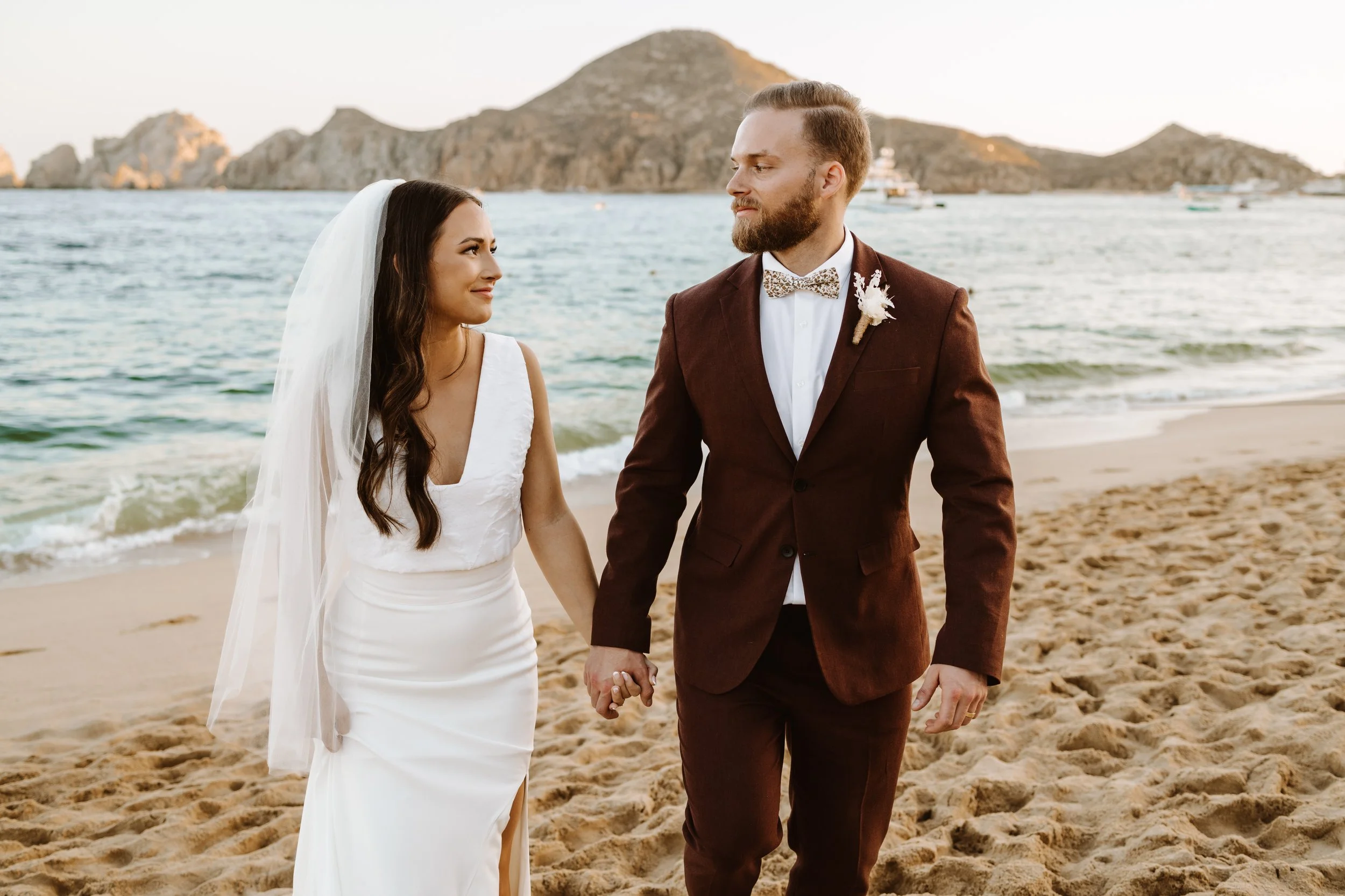 A bride and groom holding hands on a sandy beach near the ocean, with rocky islands in the background during sunset.