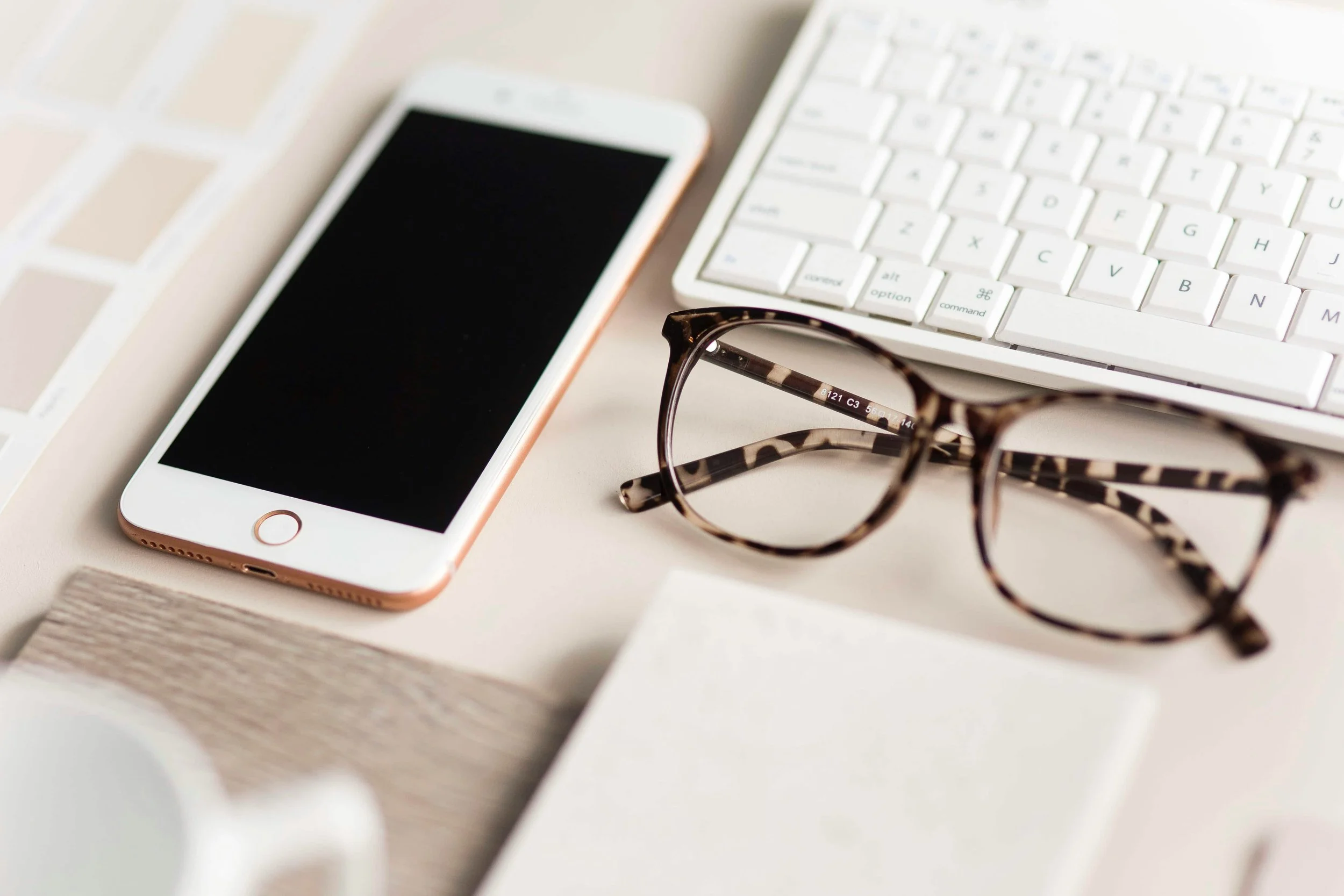 A smartphone, a pair of tortoiseshell glasses, a white computer keyboard, and a wooden surface.