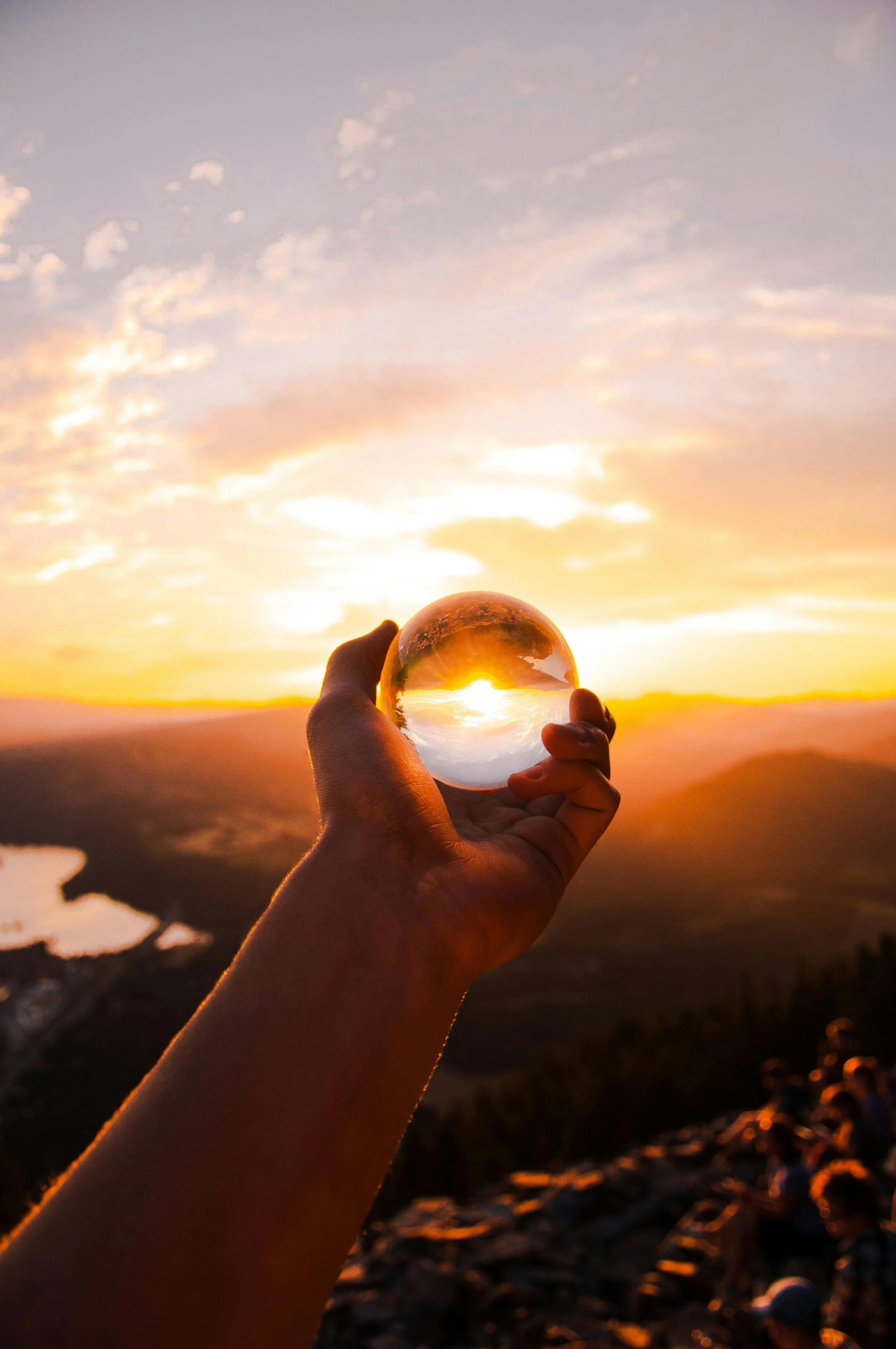 Hand holding a glass sphere reflecting a sunset over mountains and a lake.