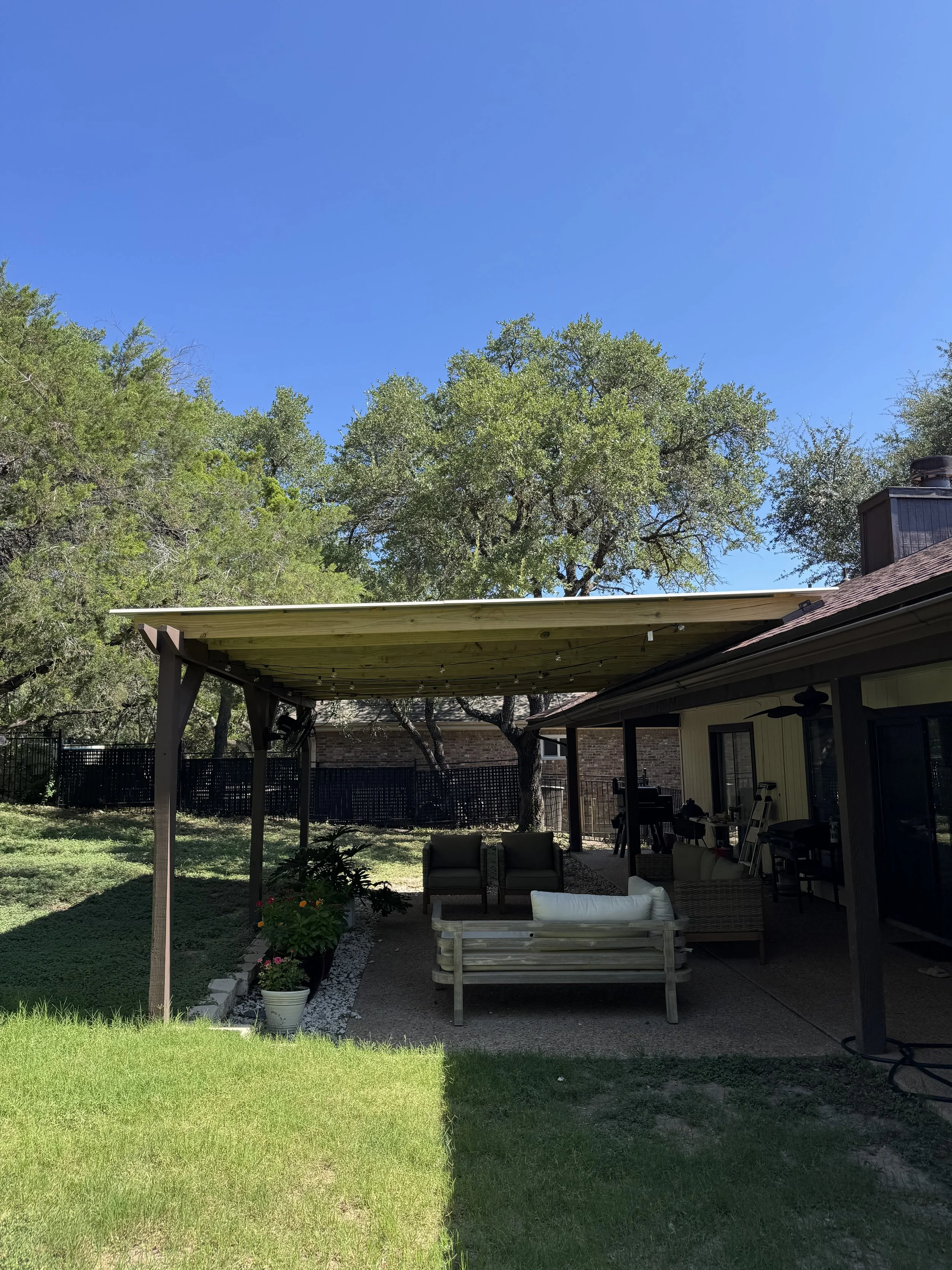 A backyard patio with outdoor furniture, potted plants, a grassy yard, trees, and a house with a covered porch underneath a bright blue sky.