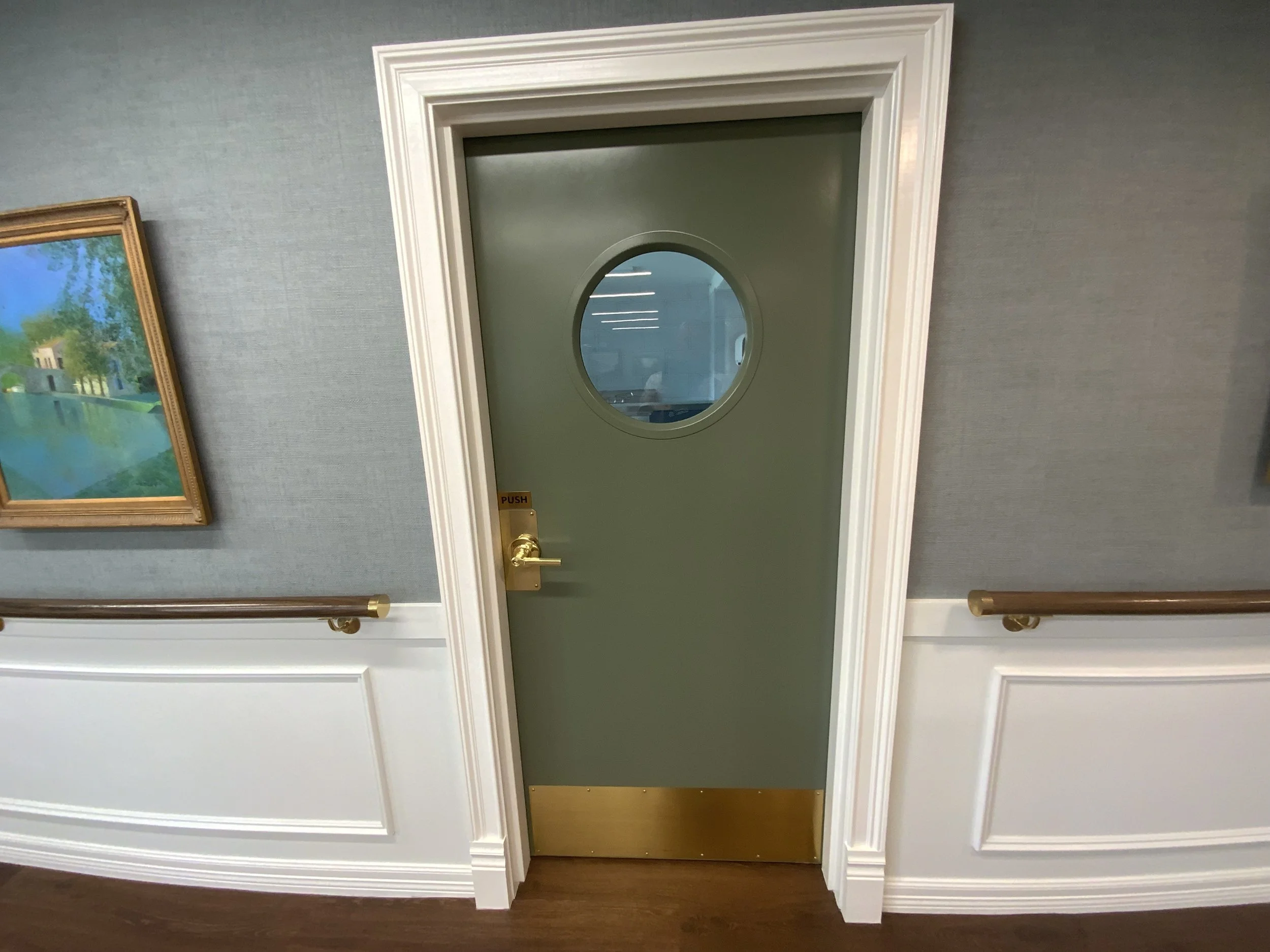 Green door with an oval window in the center, framed with white molding, set in a wall with gray textured wallpaper, next to a painting hanging on the wall.