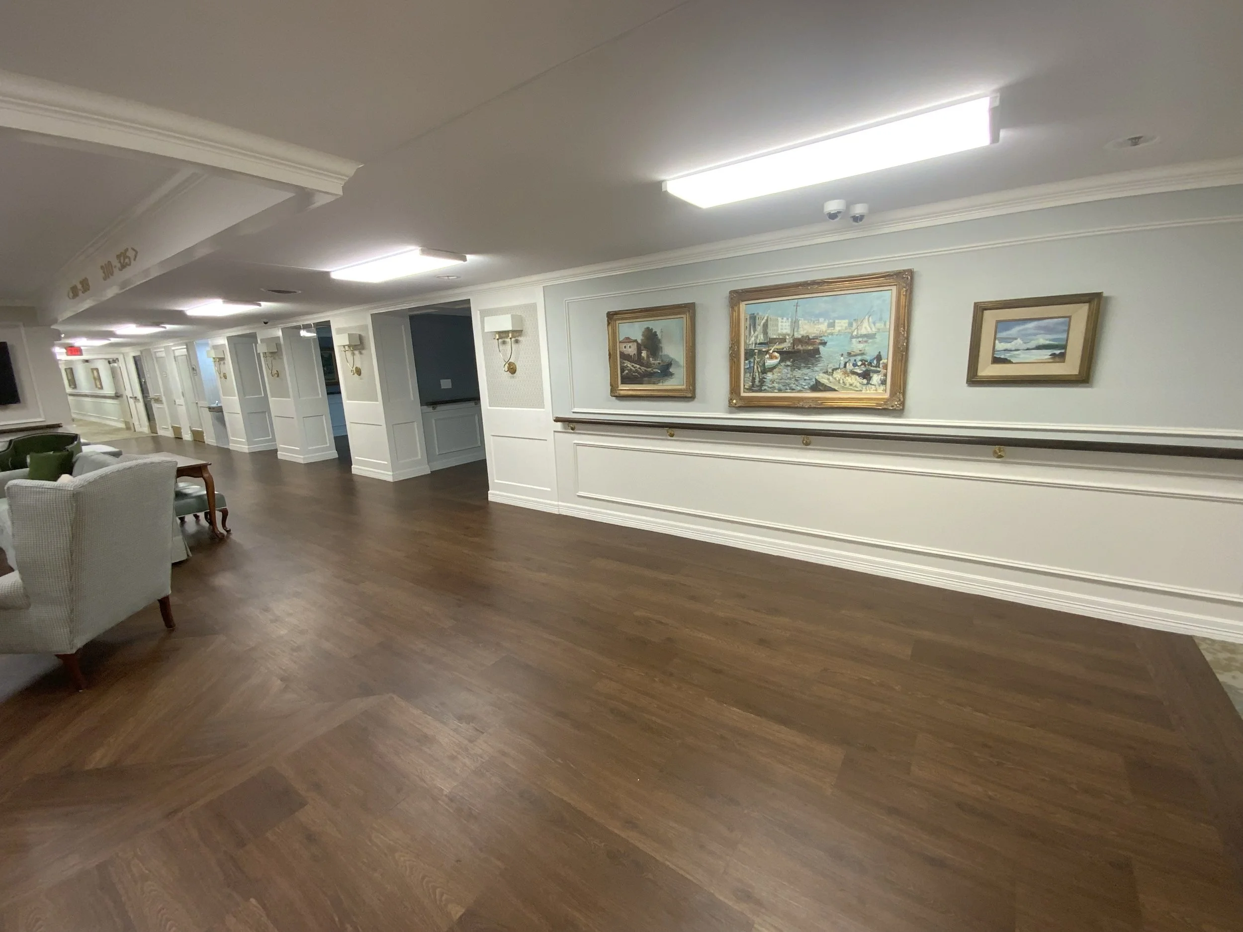 A hallway or lounge area with dark wooden floor, white walls, and paintings hanging on the wall, illuminated by bright ceiling lights.