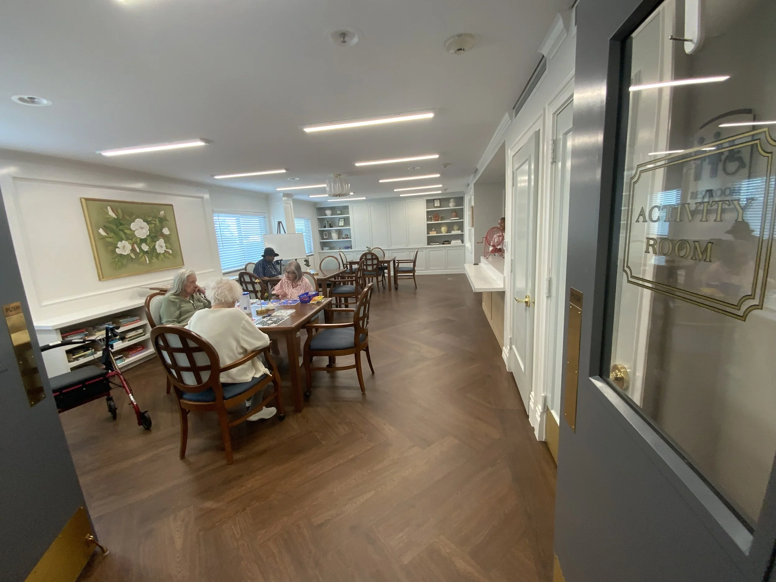 A group of elderly people sitting at a wooden table in a dining area with white cabinets and bookshelves, engaged in conversation. There is a large floral painting on the wall and windows with blinds in the background.
