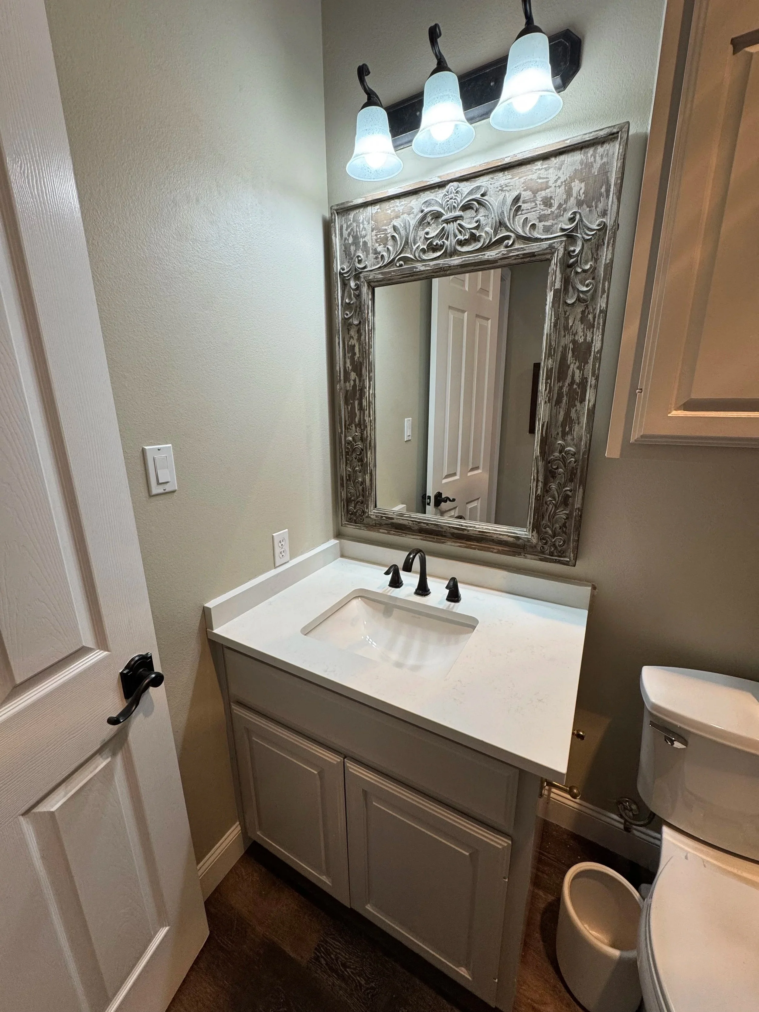 Bathroom with a white vanity, a rectangular mirror with an ornate distressed frame, and a three-light fixture above the mirror. Toilet is partially visible on the right side.