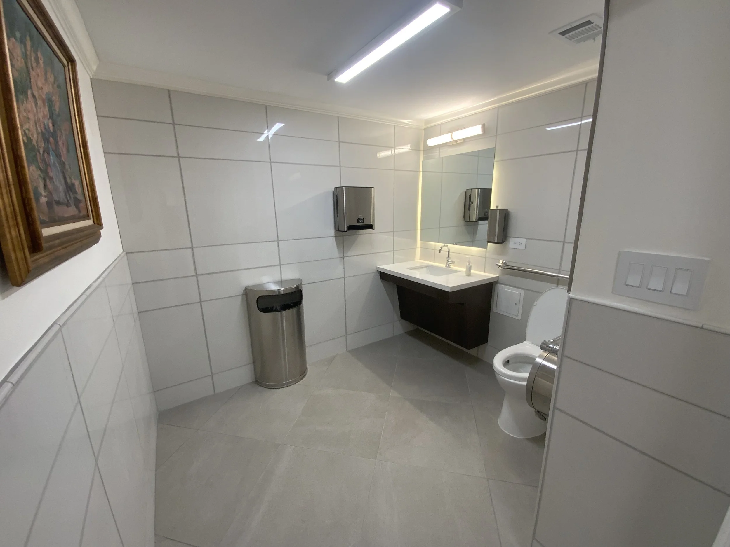 A clean and modern public restroom with white tiled walls and gray tiled floor. Features include a wall-mounted mirror with lighting, a sink with a faucet, a stainless steel soap dispenser, a paper towel dispenser, a trash can, and a toilet with a bi