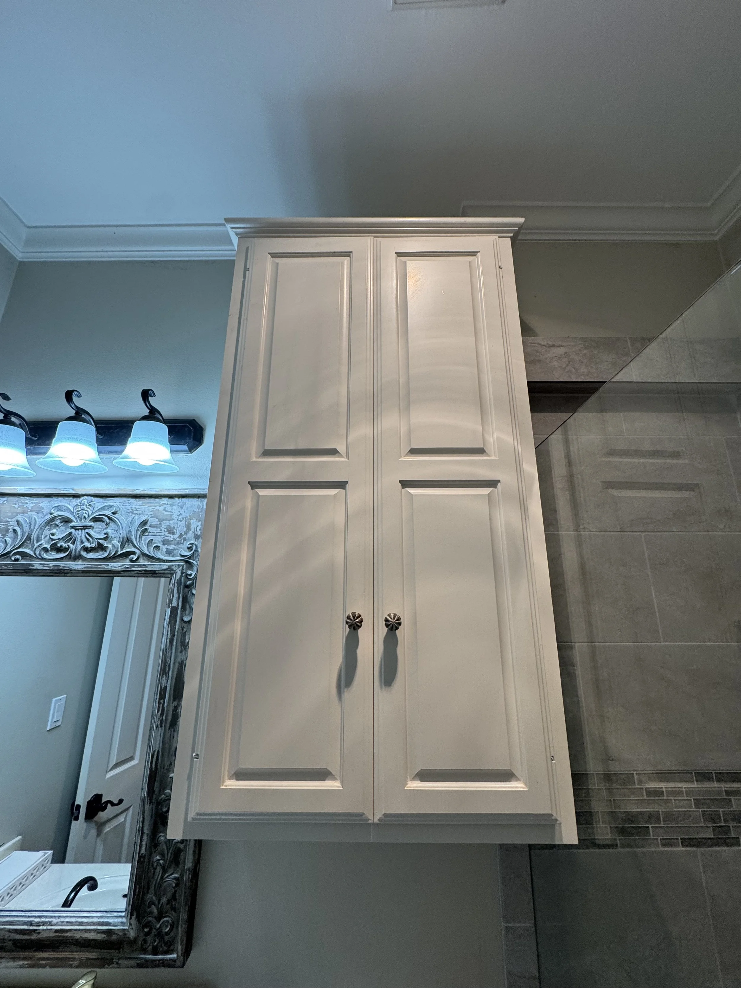 A white kitchen cabinet with two doors and round decorative knobs, positioned next to a mirror with a carved silver frame, and a wall switch visible on a light green wall. To the left, there is a section of a bathroom vanity with a black faucet and t